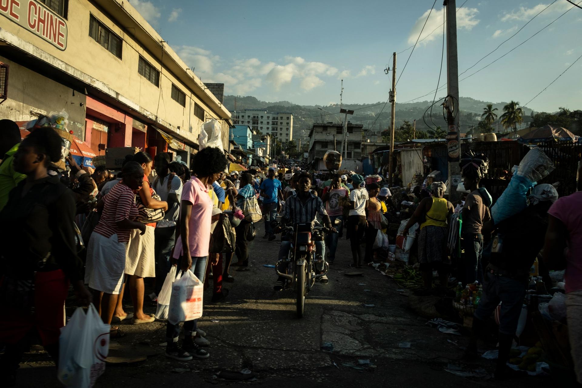 A crowd of people are shown at a Haitian market in Port-au-Prince with a person on a motorcycle in the middle.