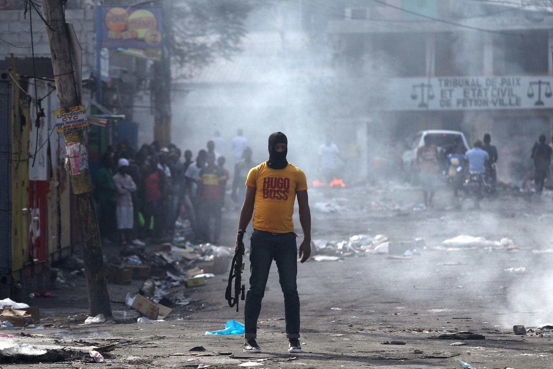 A man is shown holding a weapon, wearing a yellow Hugo Boss t-shirt, next to burning barricades.