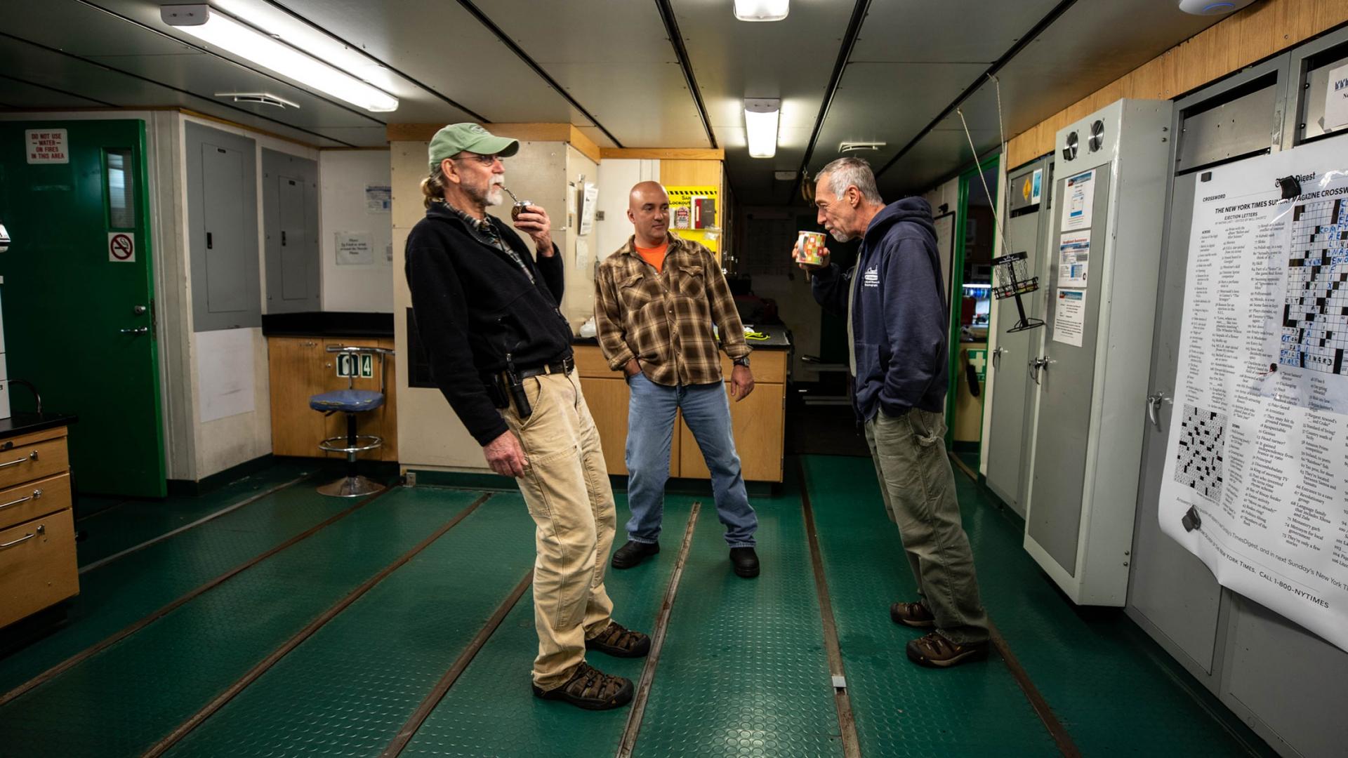 Three crew members are shown in a room with a green floor all leaning in order to stay balanced.