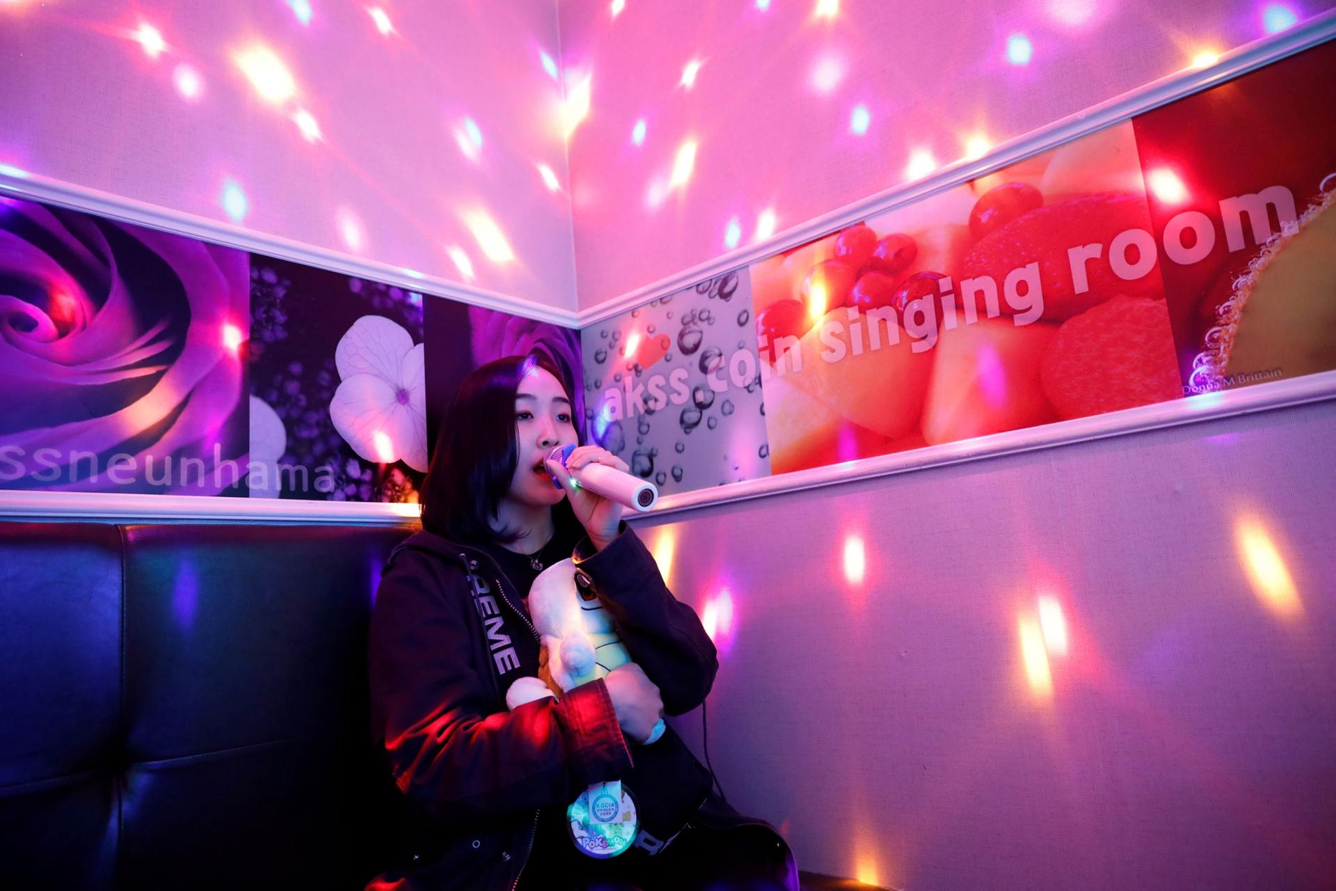 A woman is shown singing into a microphone with pink lights on the walls.