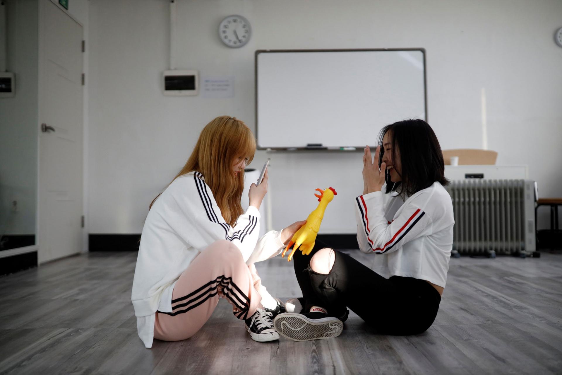 Two young women are shown sitting on the floor with one holding a yellow rubber chicken and taking a photo with her mobile phone.