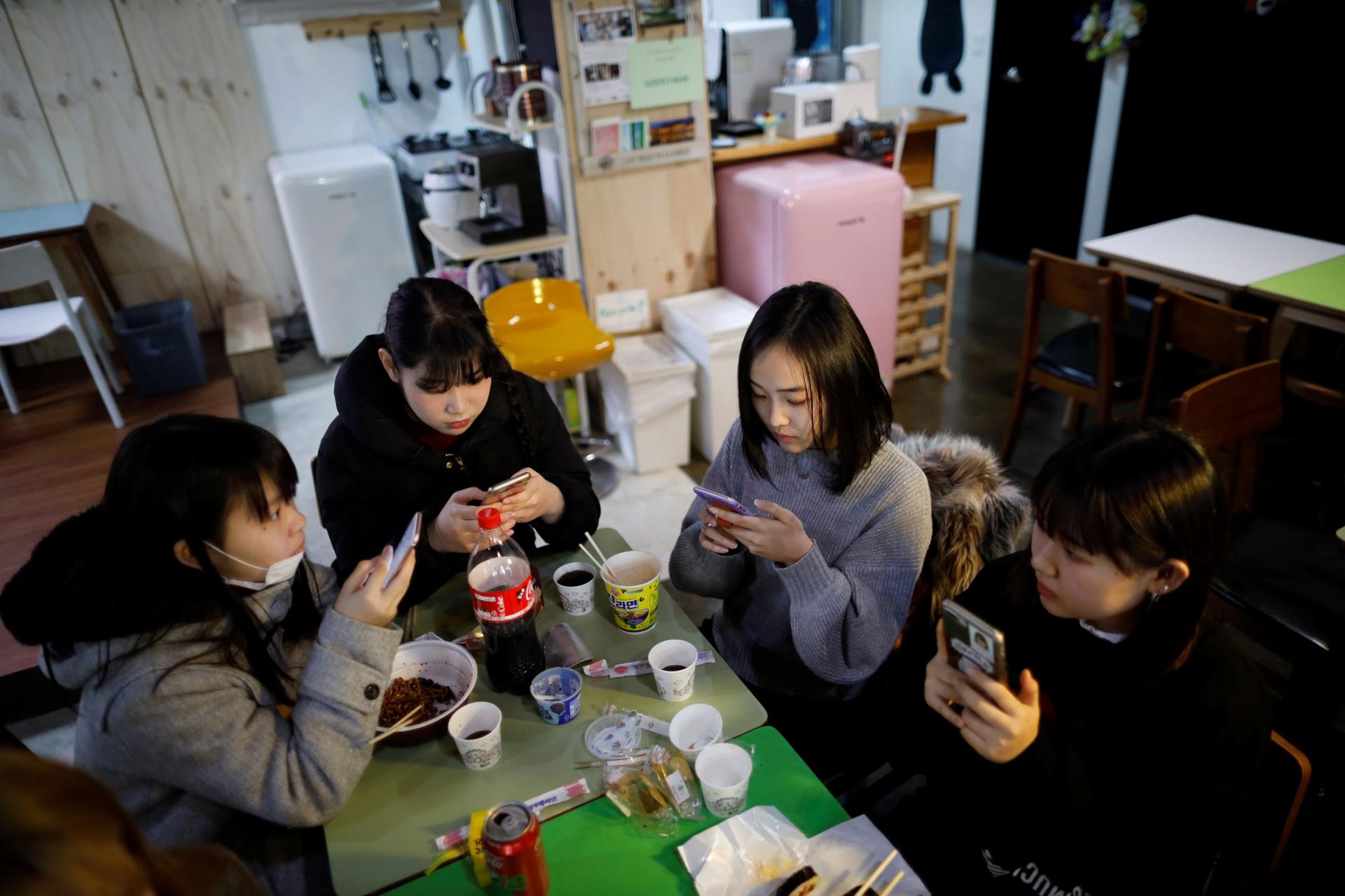Four young women are shown sitting around two small green tables, all of them with a mobile phone.