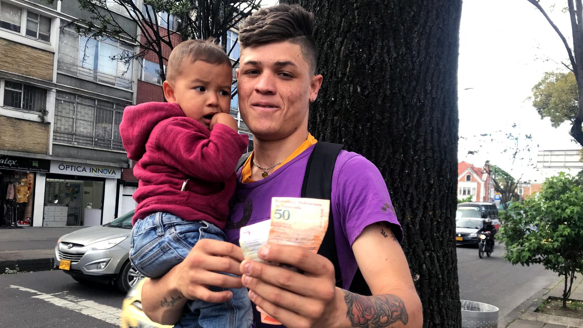 A young man holds a baby boy in his arms and hands out worthless Venezuelan bolivar. 