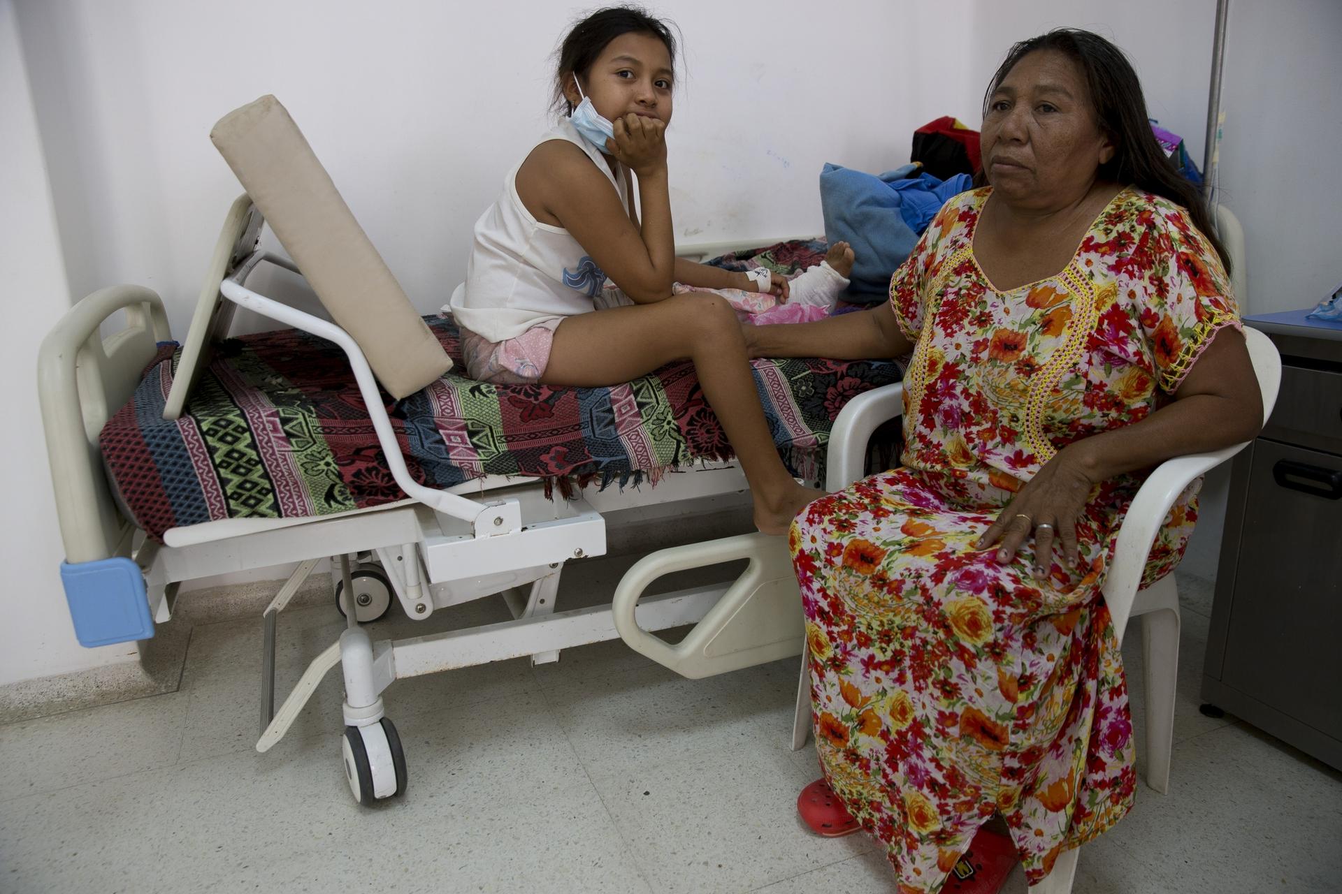 A young girl sits on a hospital bed with a fractured leg in bandages next to her mother. 