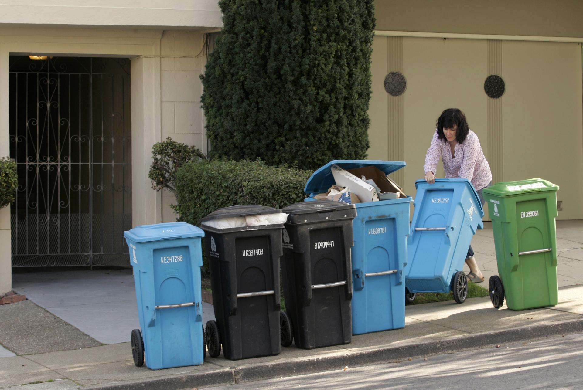 A woman puts different large garbage and recycling containers on a sidewalk. 