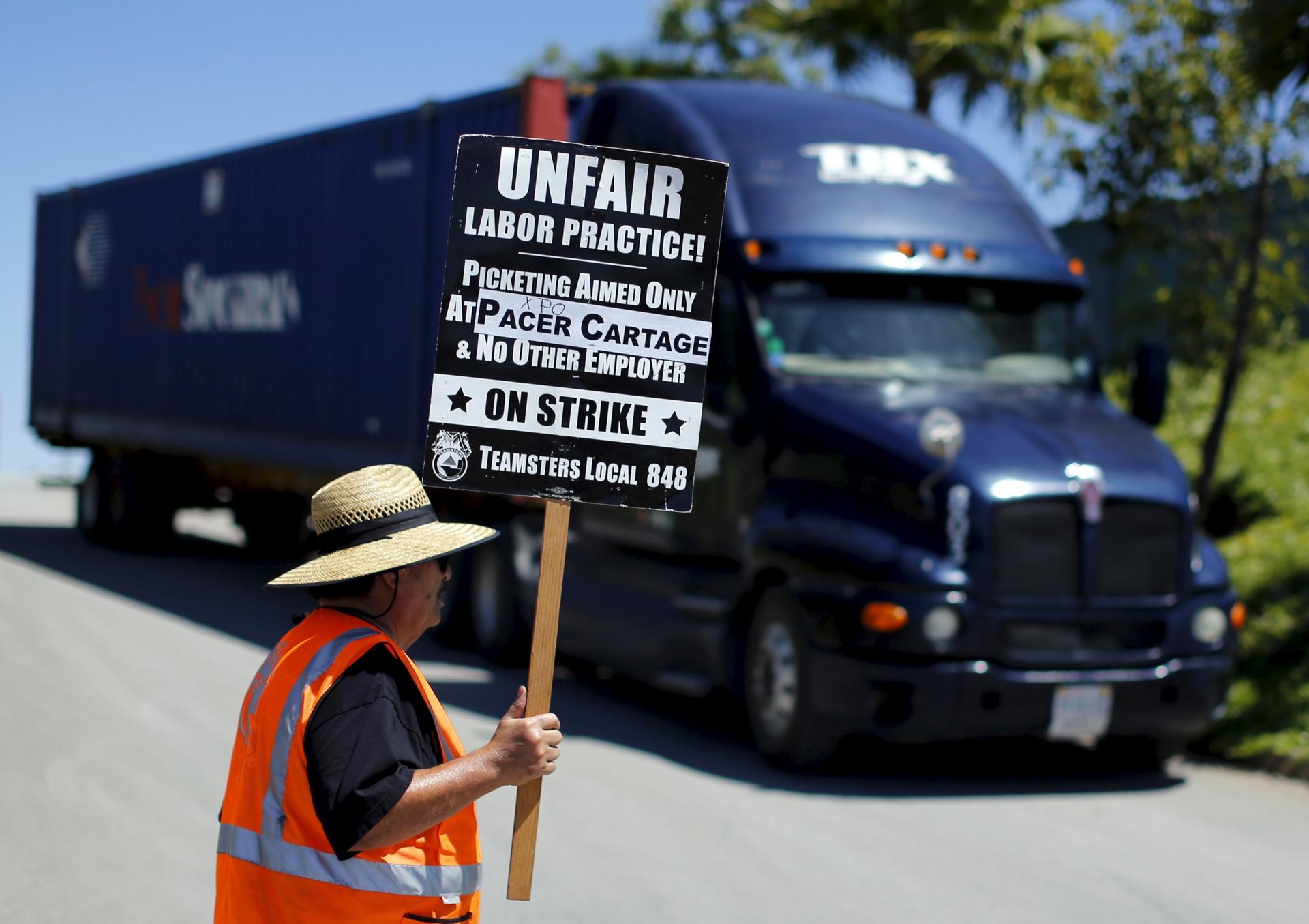 A man next to a truck stands in an orange vest with a sign that says "unfair labor practice."