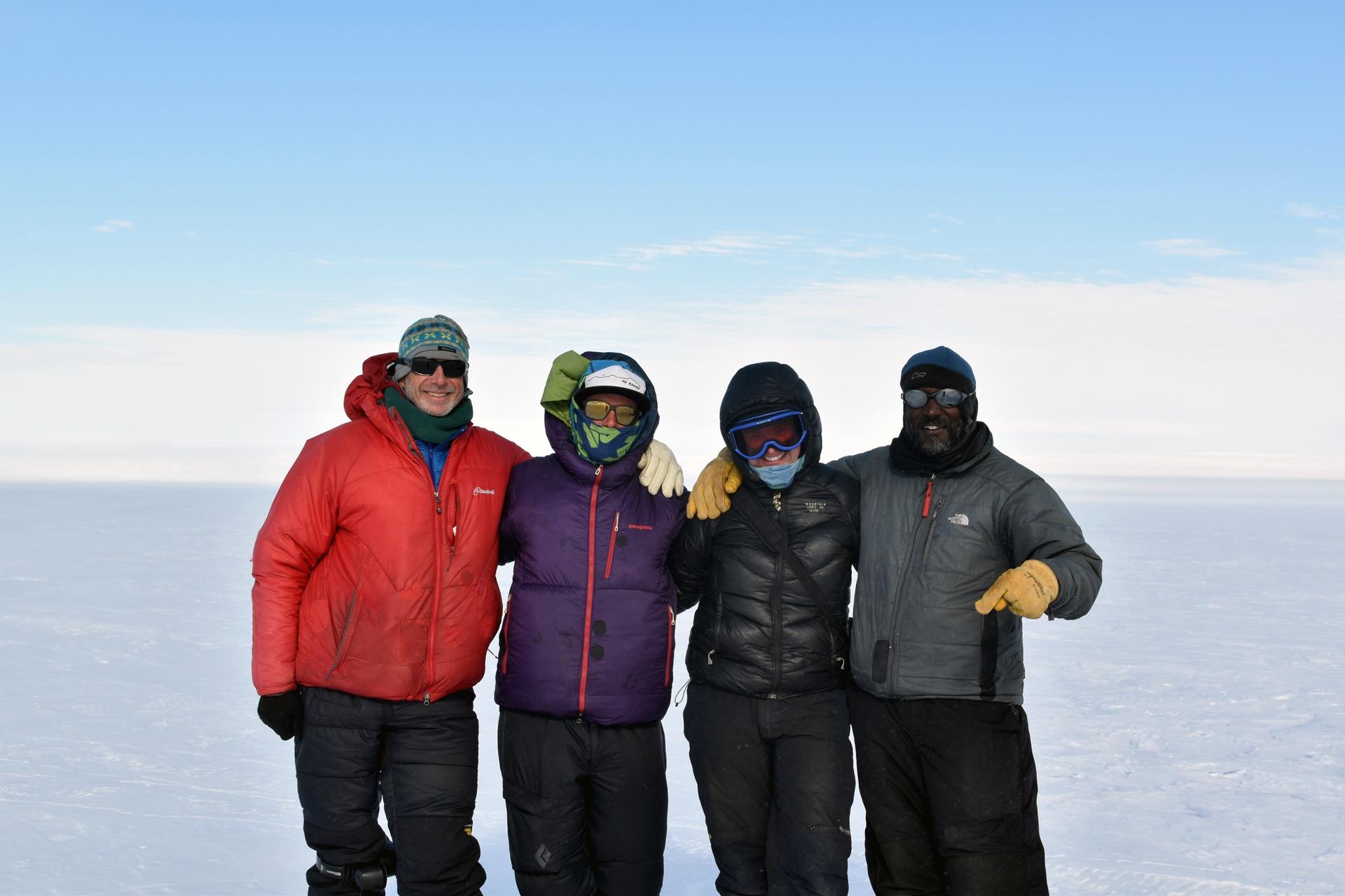 A group of four people pose in cold weather gear. Behind them is a white, icy horizon.