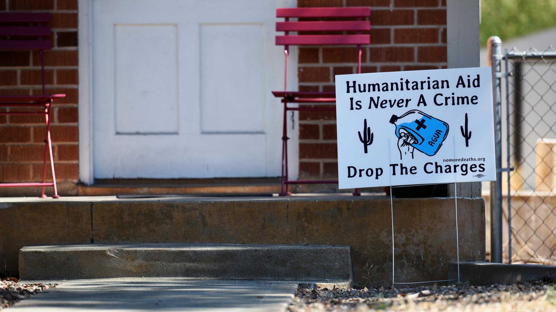 A yard sign says "Humanitarian aid is never a crime. Drop the charges." and includes a picture of a waterbottle and cactus. 