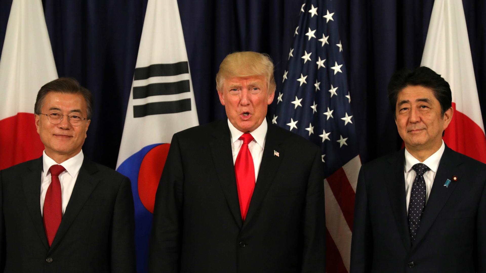South Korean President Moon Jae-in, US President Donald Trump and Japanese Shinzō Abe stand in a line with flags form their countries behind them.