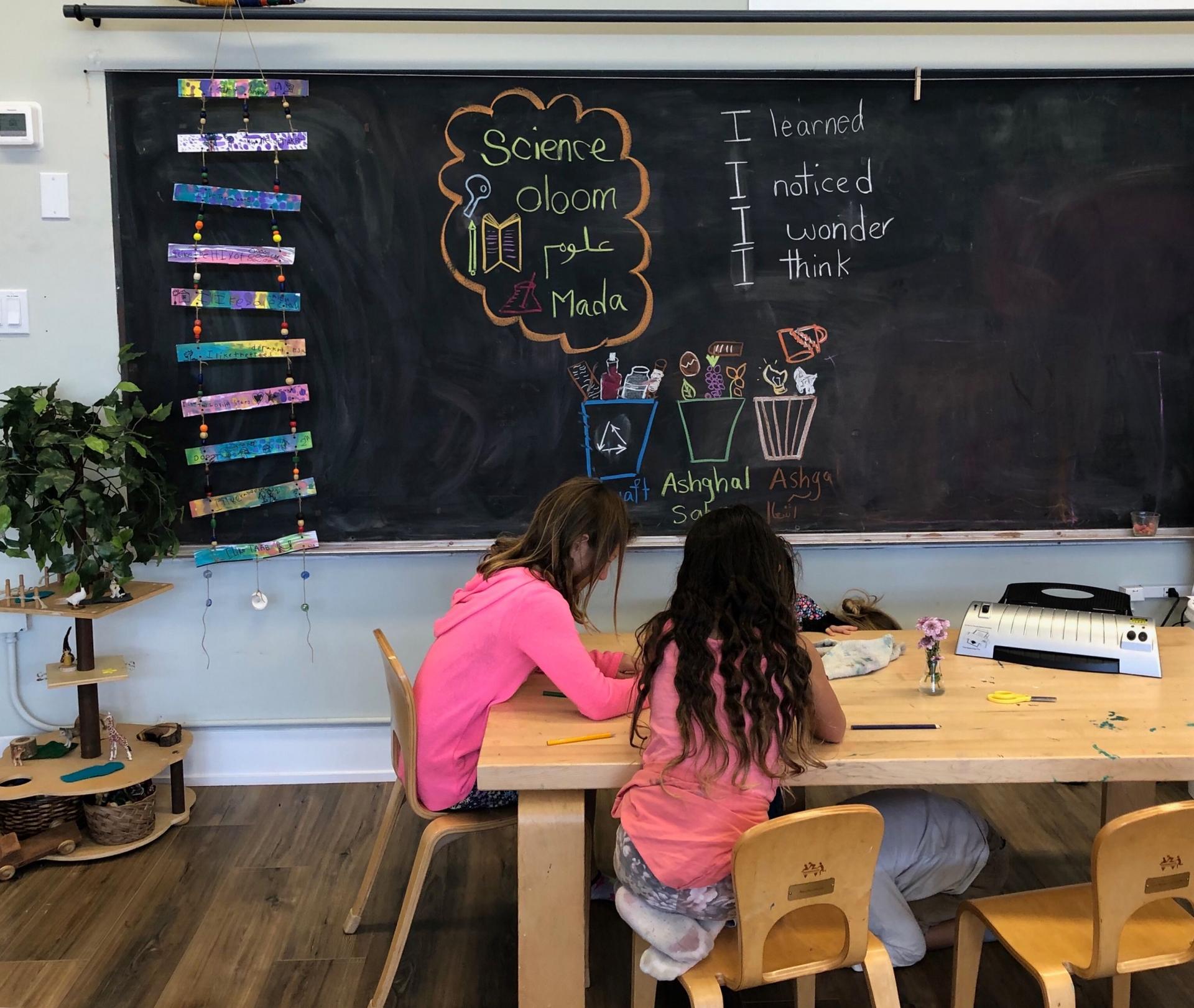 Two young girls sit a table in front of a chalkboard. 