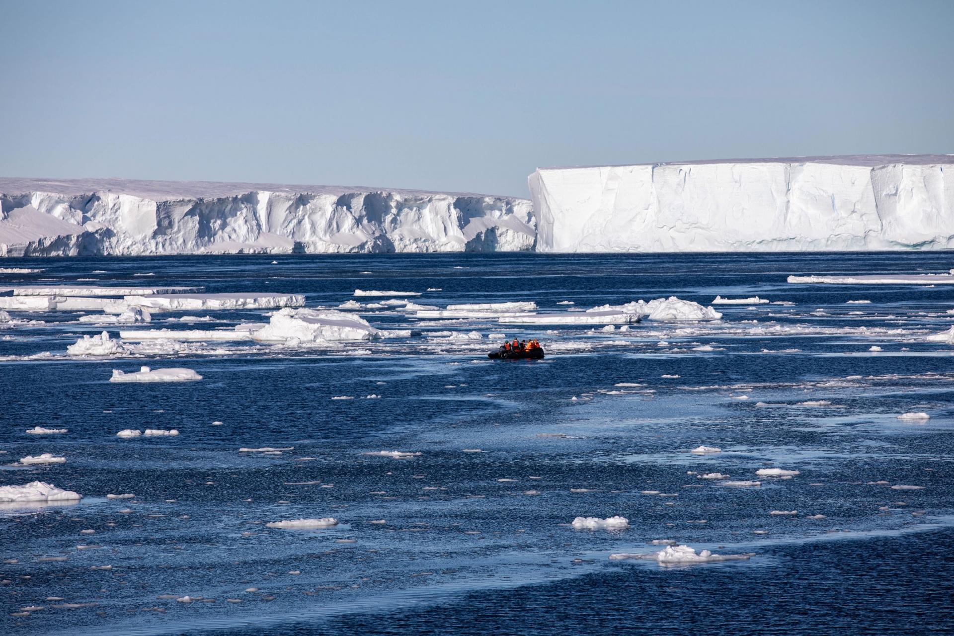A Zodiac boat is shown off in the distance with several people aboard with large white floating ice all around.
