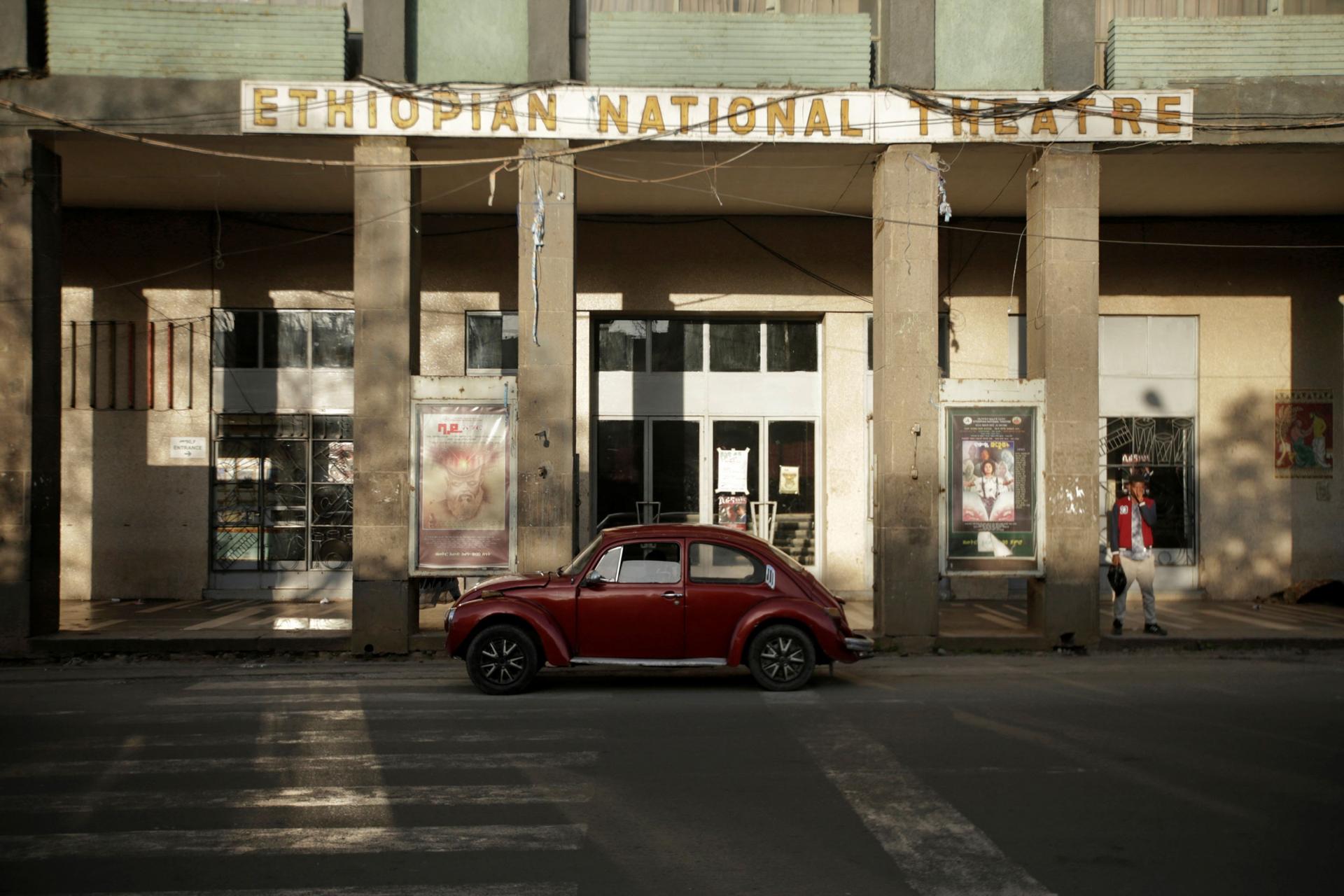 A red Volkswagen Beetle is show parked in front of the tall columns of the Ethiopian National Theatre.