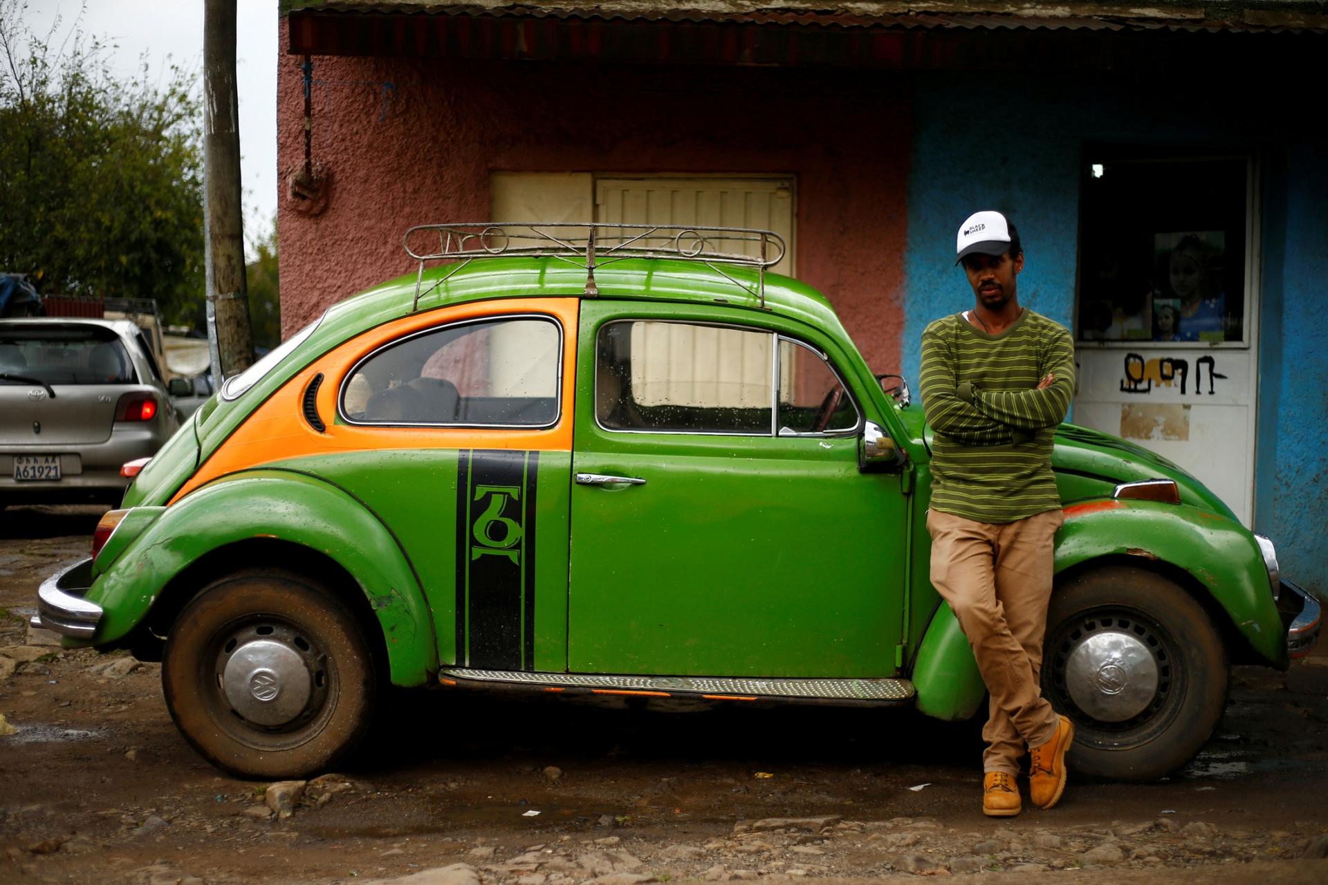 A man in a stripped shirt and hat is shown standing next to a green 1976 model Volkswagen Beetle.