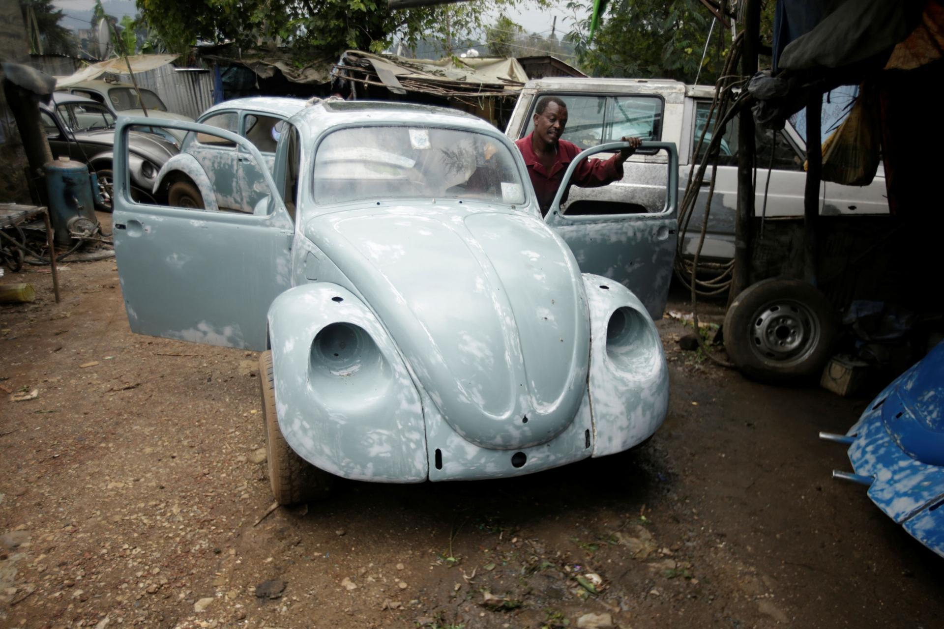 A man is shown behind an open door of a Volkswagen Beetle while he is pushing it.