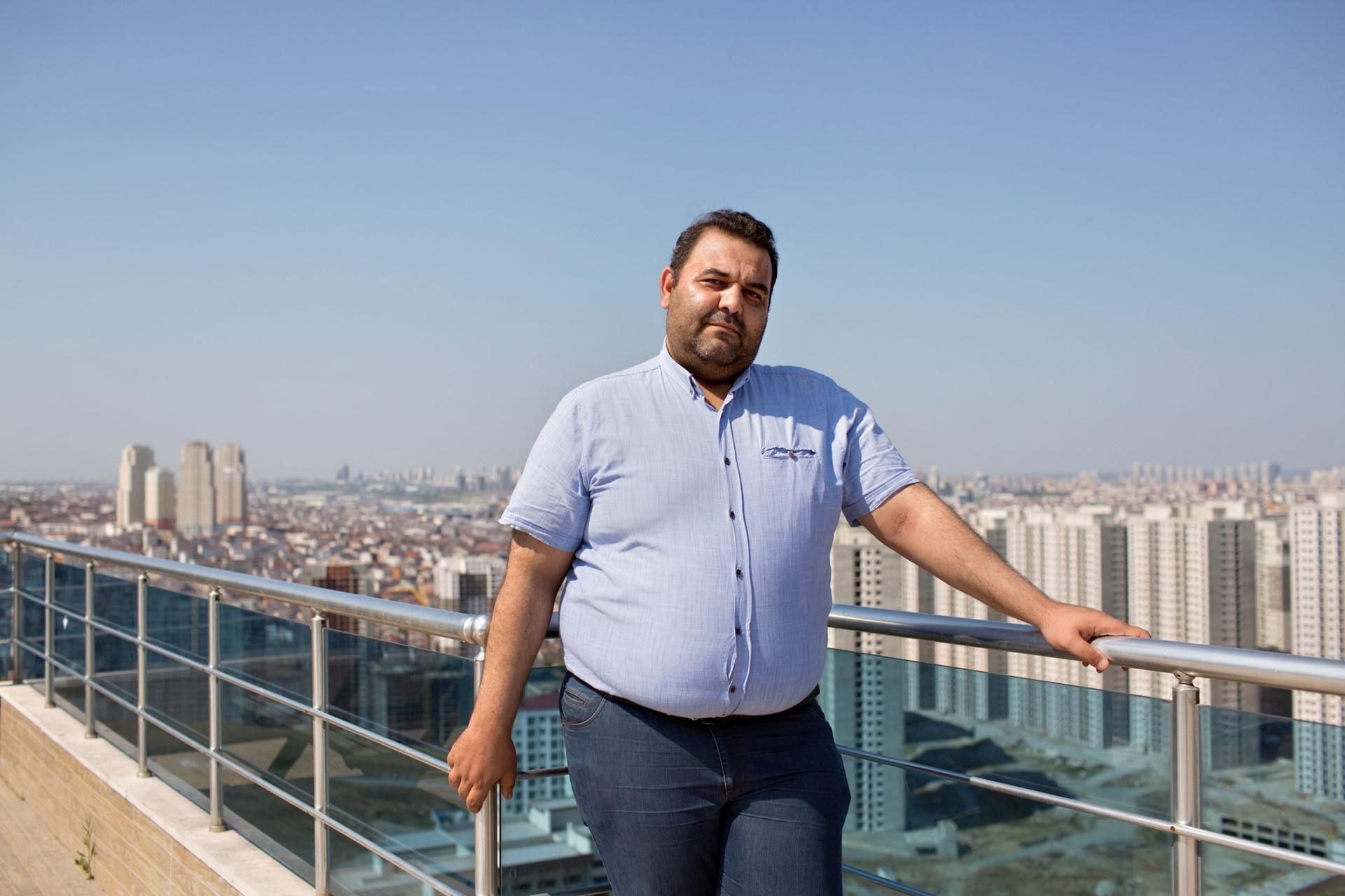 A man is shown in a blue button down shirt and jeans leaning against the railing of a rooftop deck.