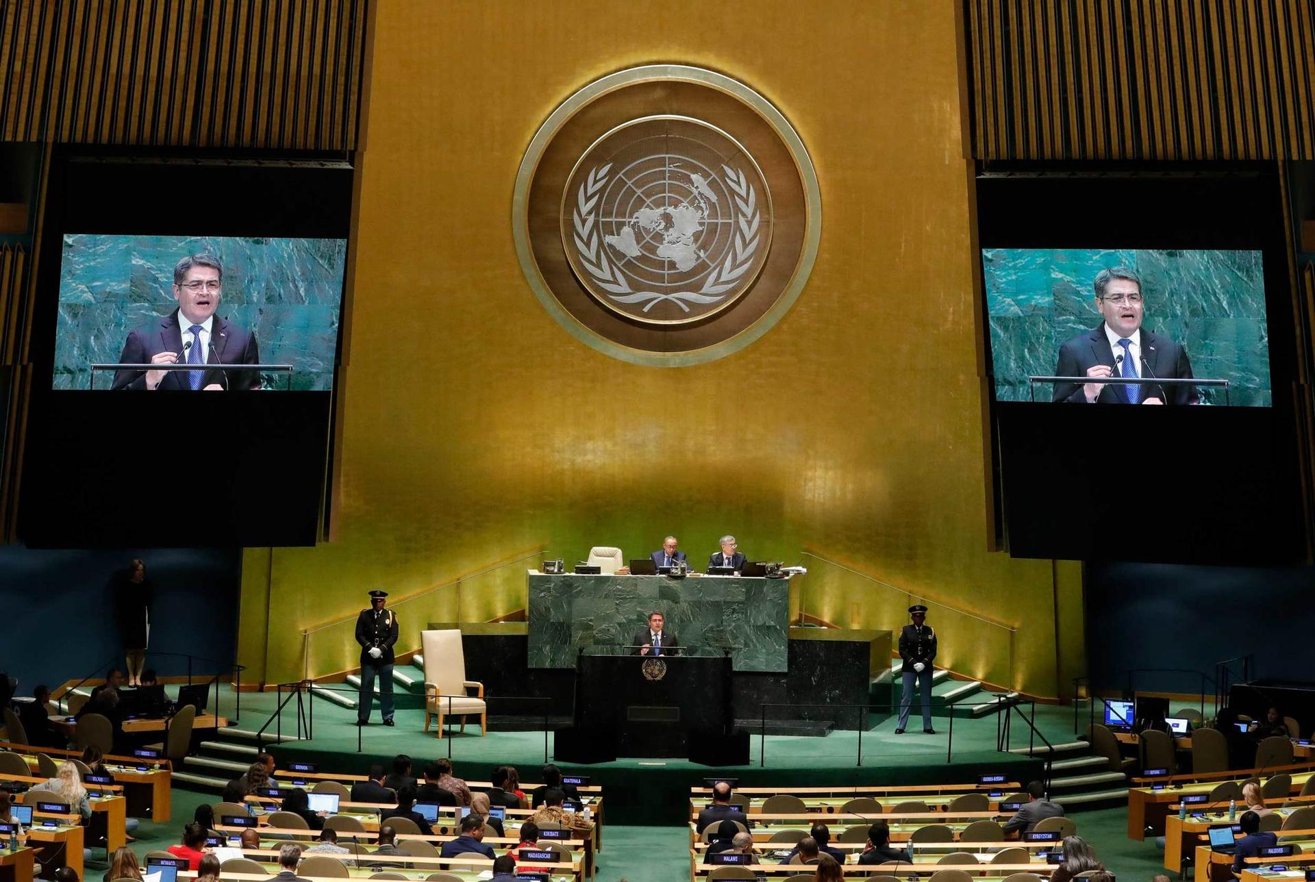 A man speaks at the UN. His image is seen in two screens at left and right of the stage.