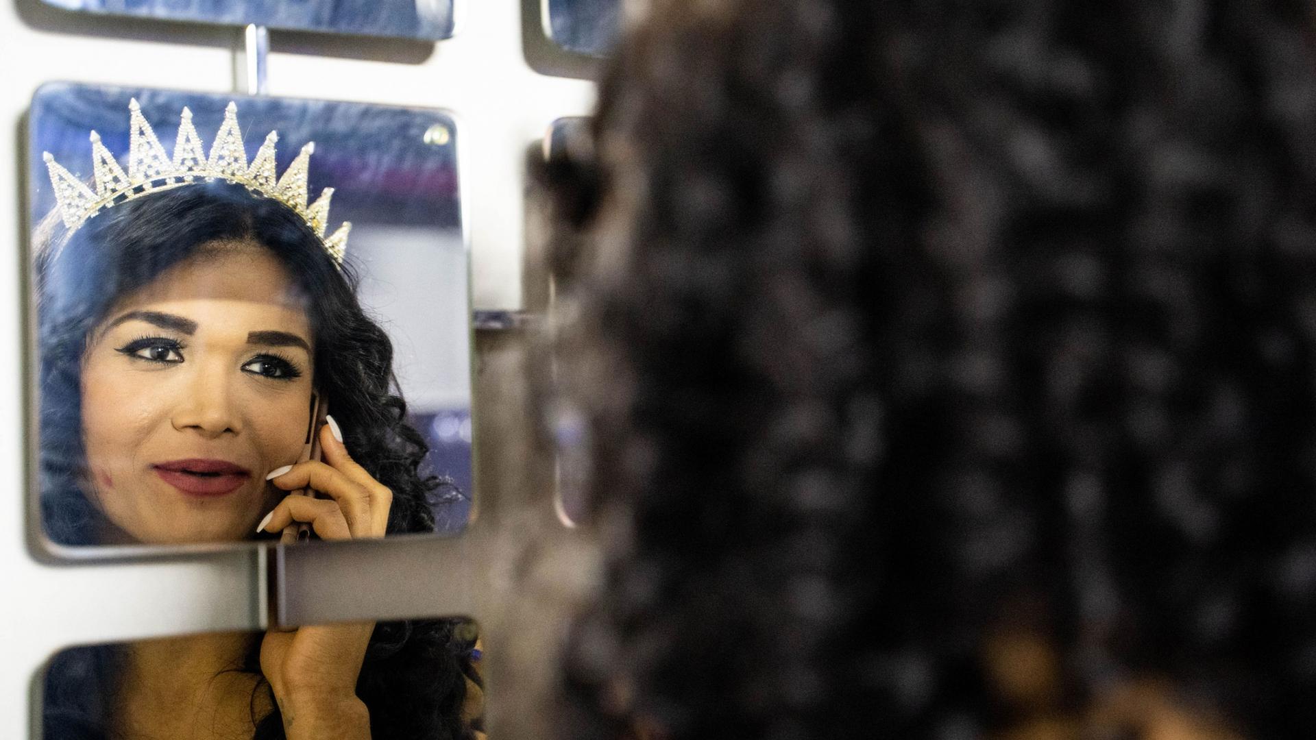 A transgender woman is shown in the reflection of a mirror holding a phone to her hear and wearing a crown.