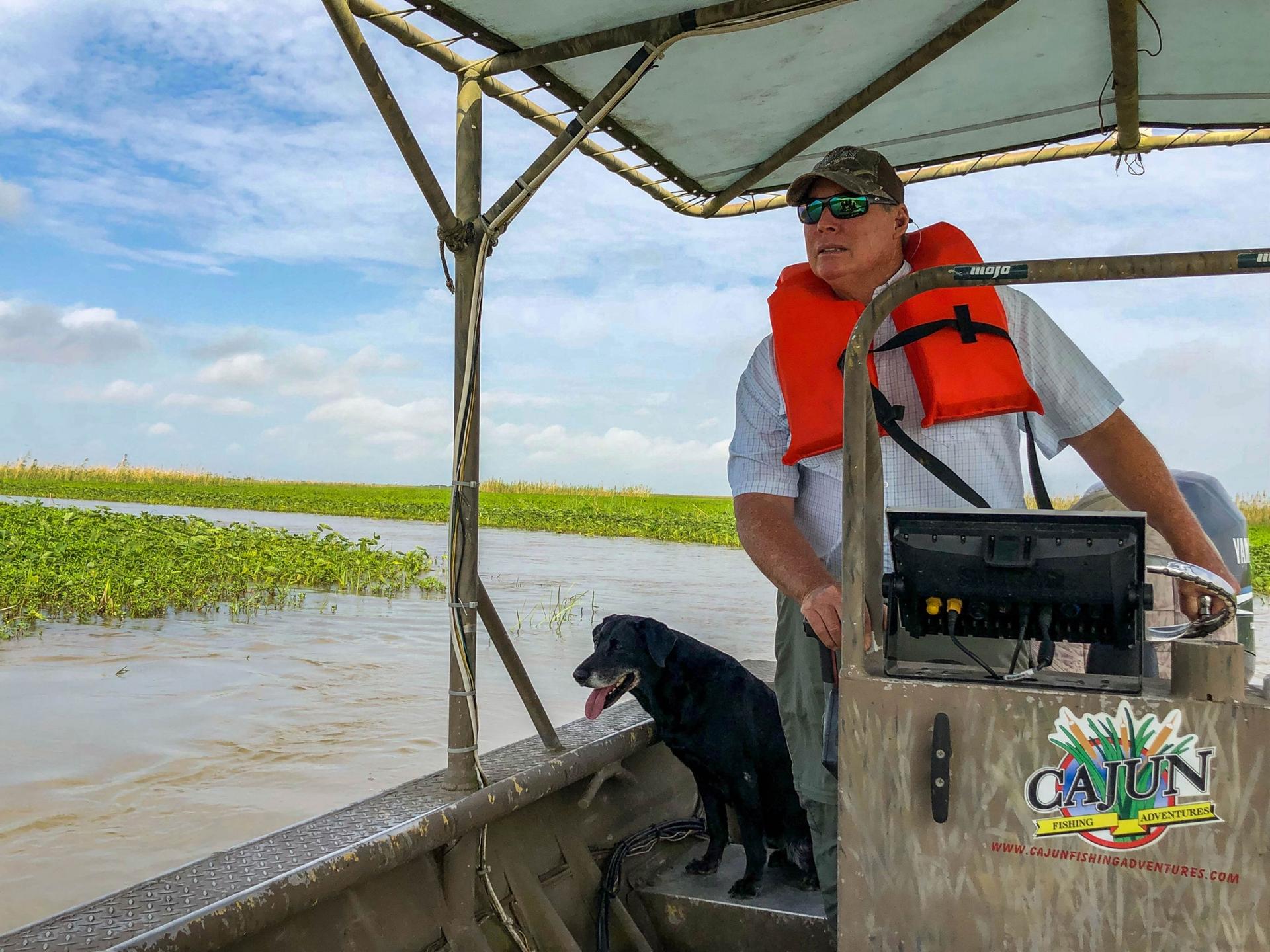 Ryan Lambert is shown piloting a flat bottom boat with a dark colored dog sitting next to him.
