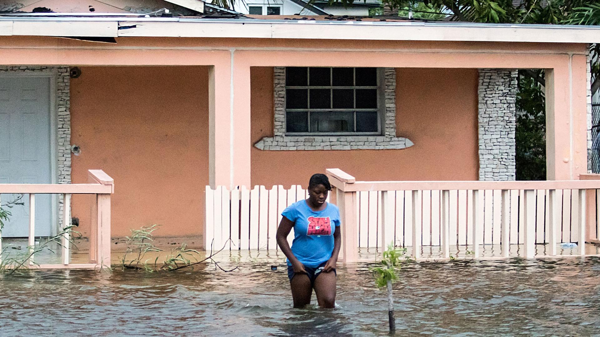 A woman is shown walking in knee-deep water in front of a peach-colored home.