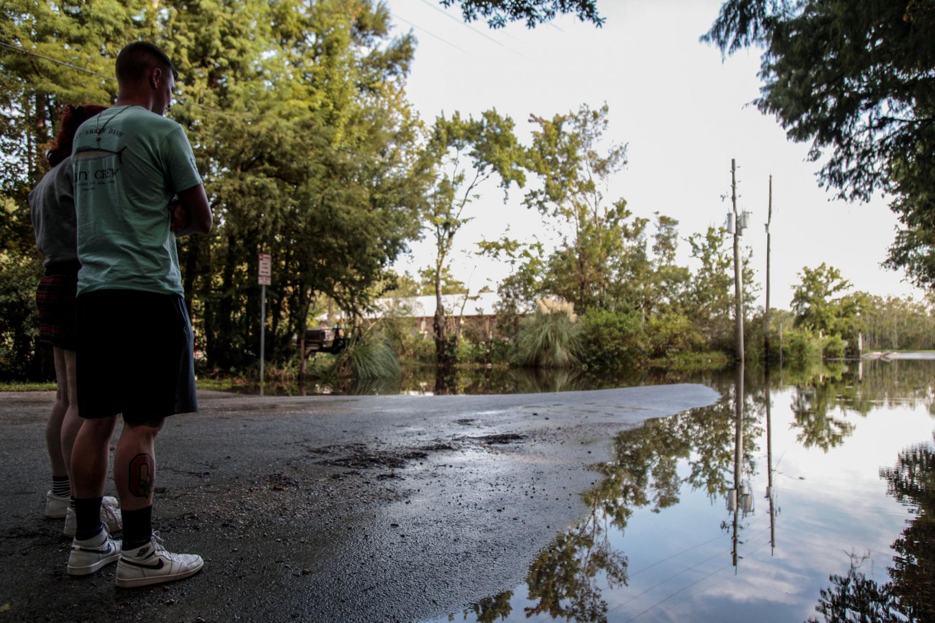 Two people are shown standing on a tree-lined street partially flooded from recent storms.