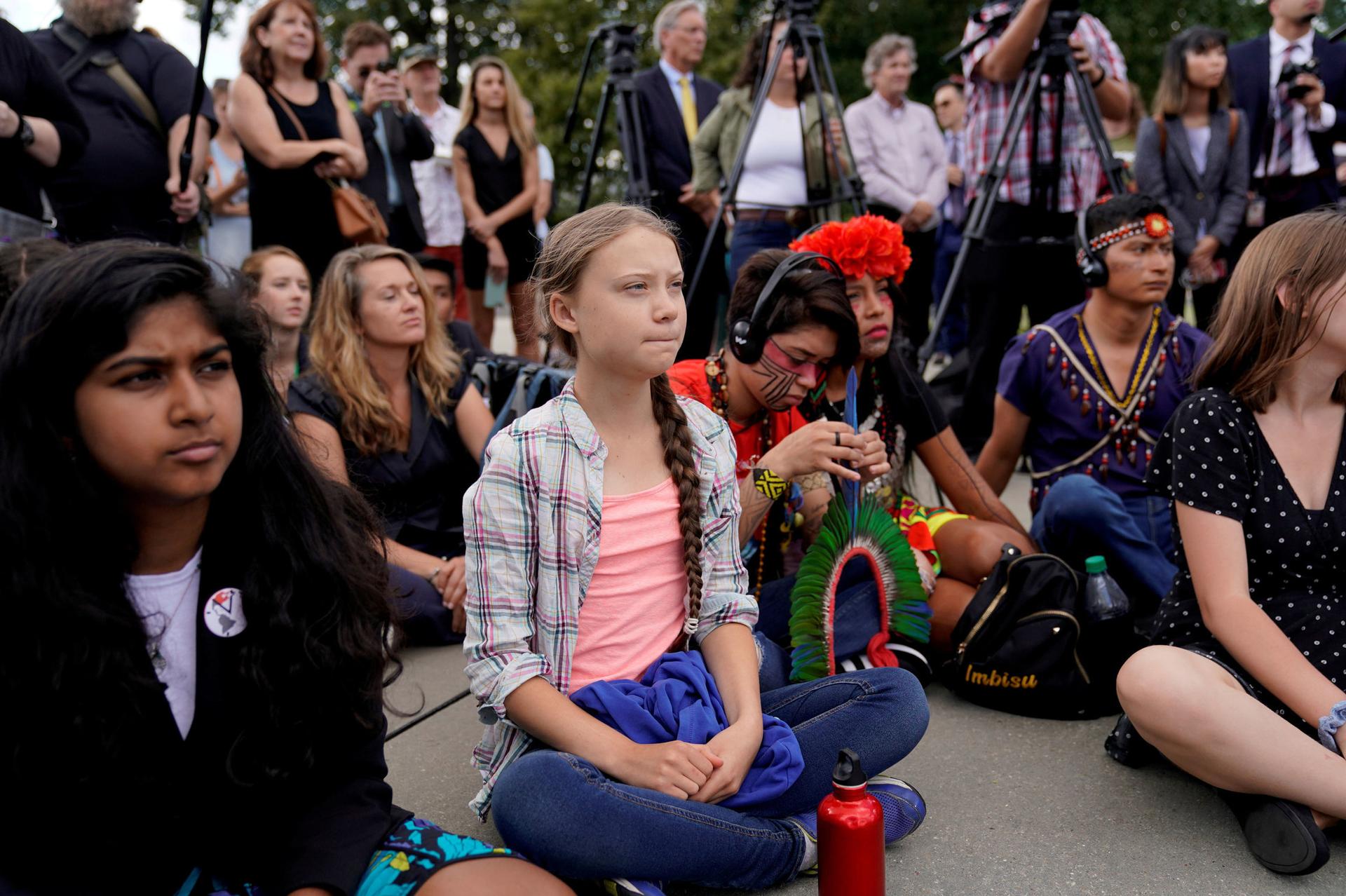 Swedish climate activist Greta Thunberg is shown sitting on the ground with her legs crossed and with a red water bottle.