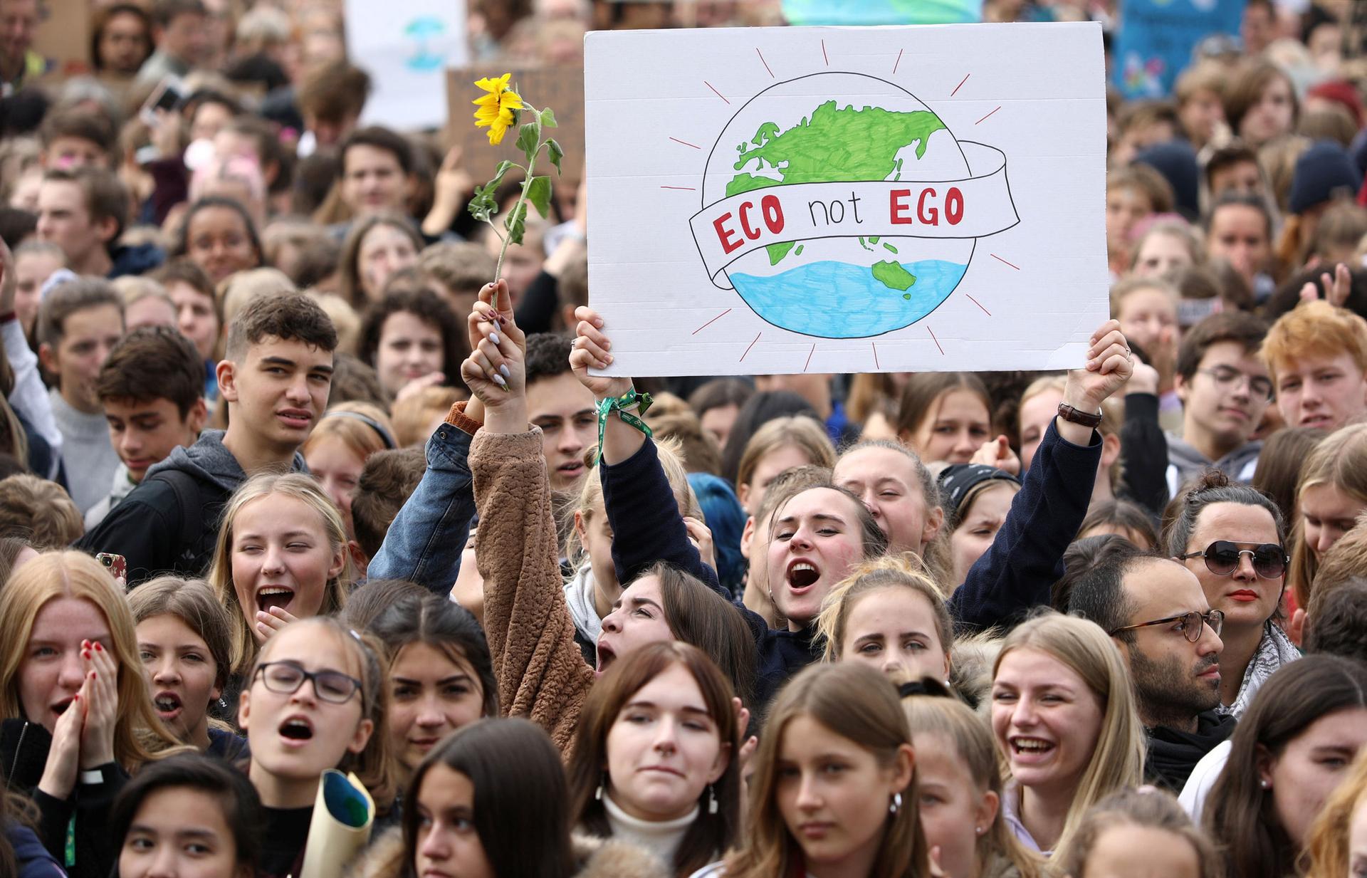 A large crowd of people are shown with one woman hollding a sign that reads, "Eco not Ego."