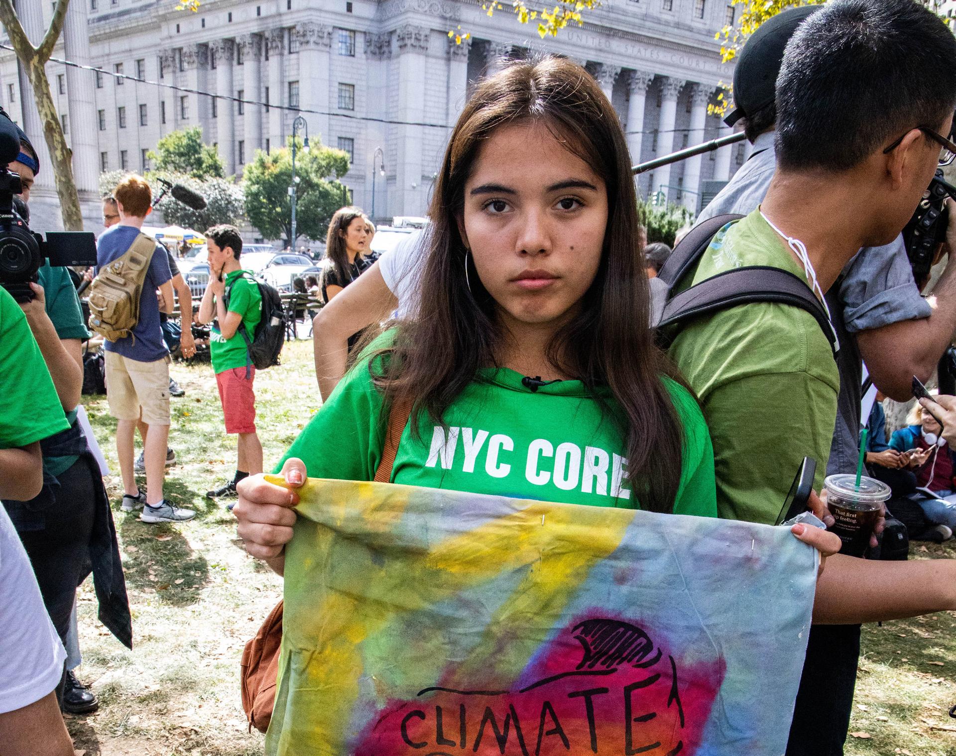 Xiye Bastida is shown standing with a green sweatshirt on and holding a sign with the word, "Climate," written on it.