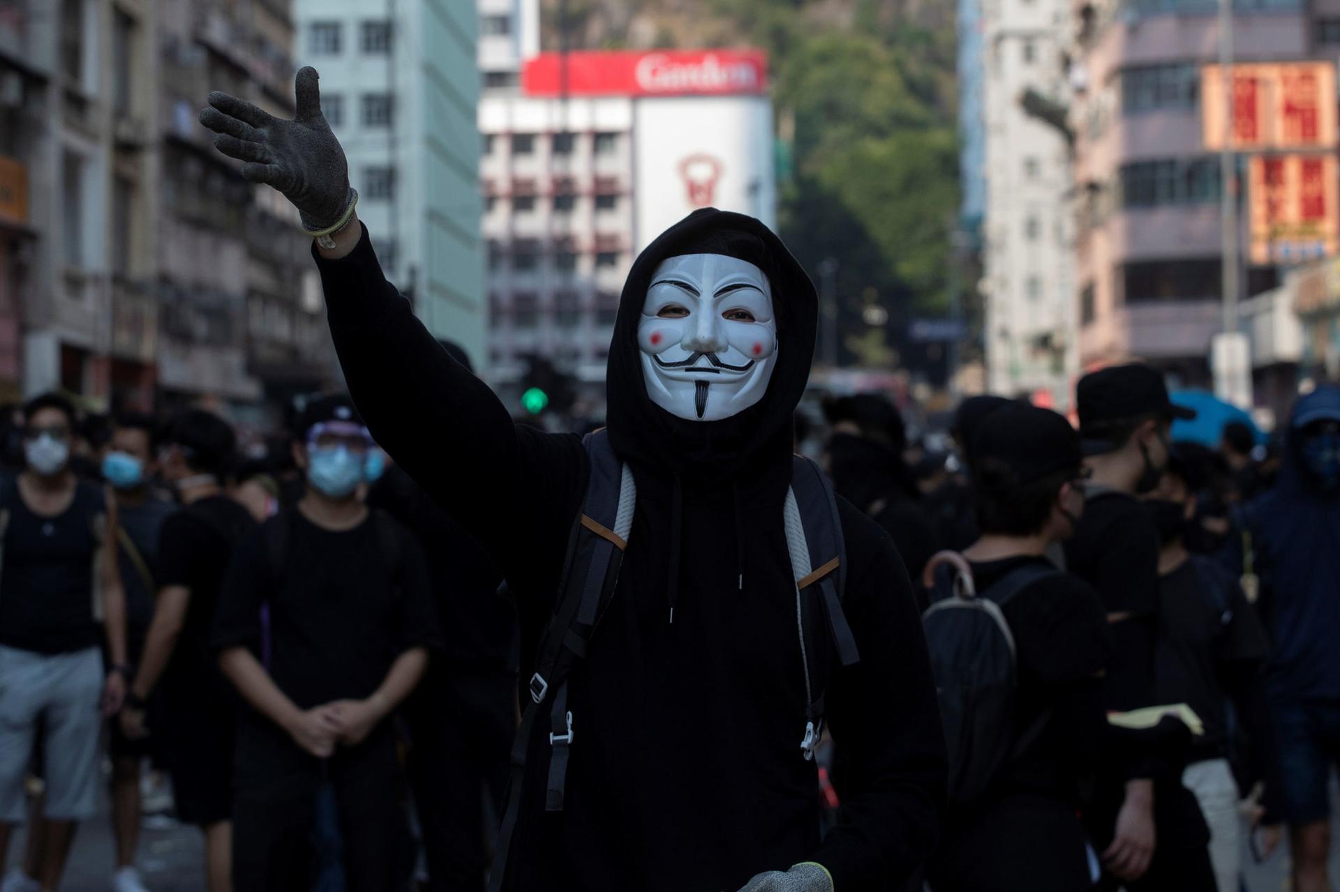 A person is shown center frame wearing a Guy Fawkes maks with their right raised hand in the air.