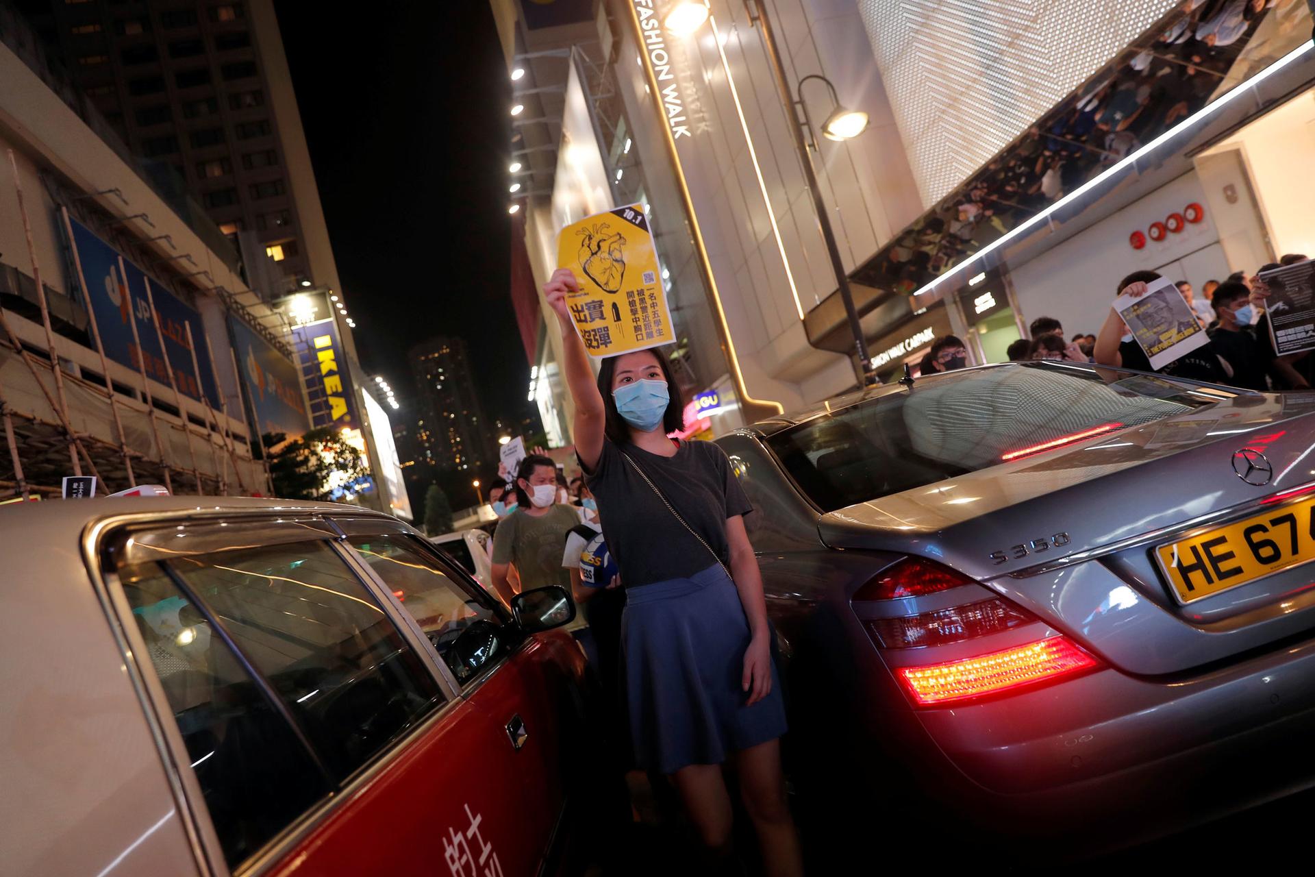 A woman is shown wearing a hospital mask and standing inbetween two cars.