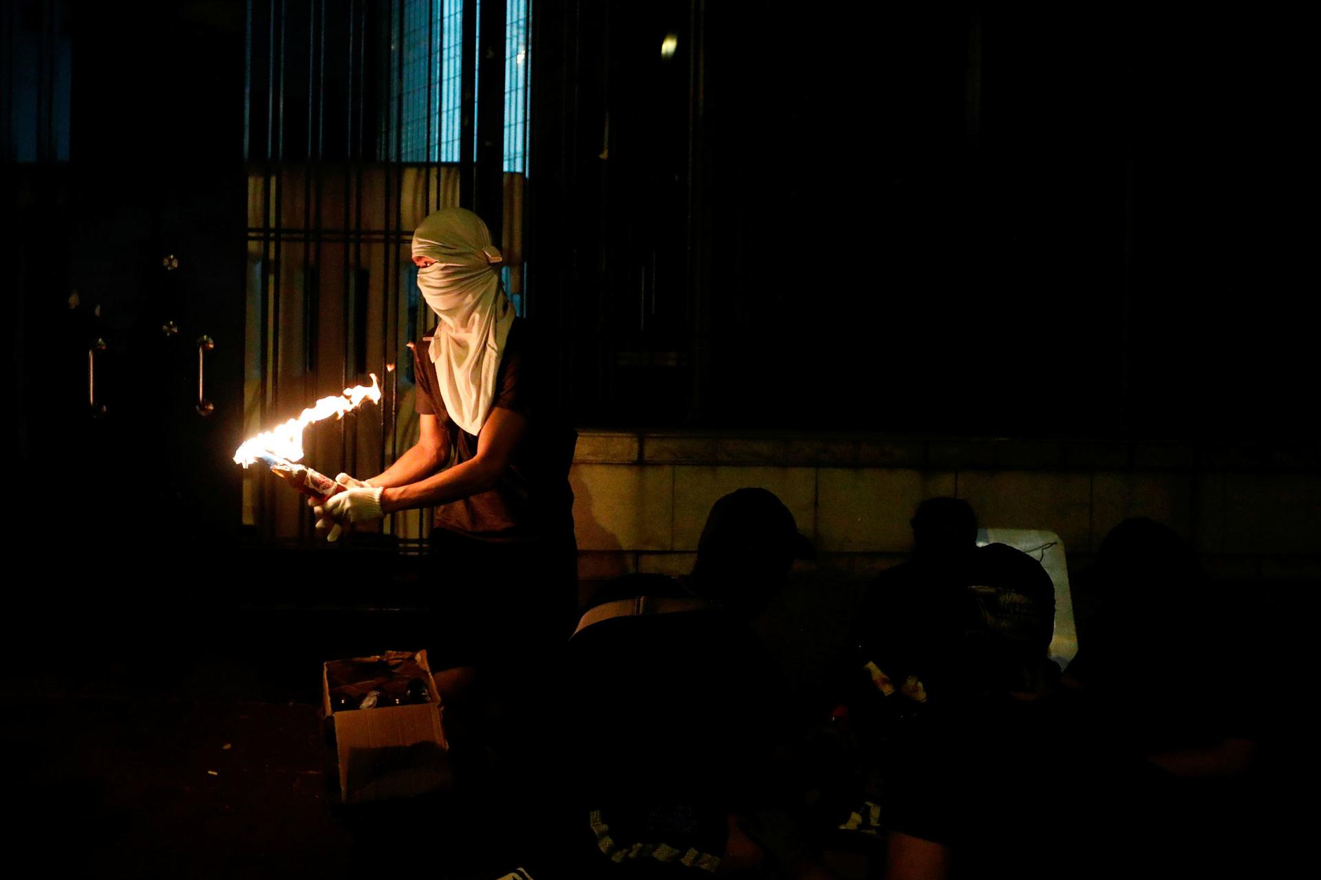 An anti-government protester is shown holding a Molotov cocktail set ablaze and wearing white mask covering his face and head.