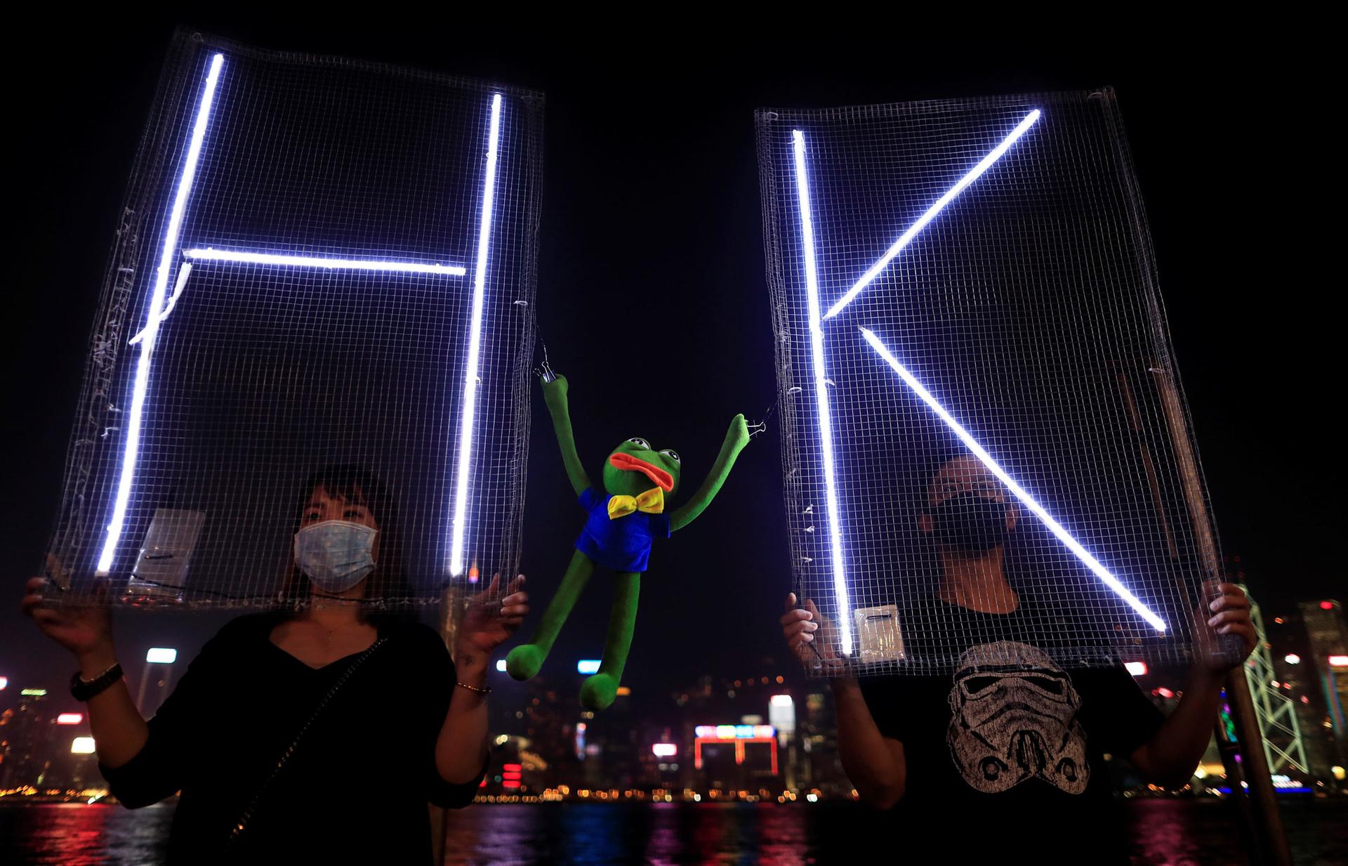 Two people are shown holding signs with electrified letters H and K with a Pepe the Frog plush toy inbetween.