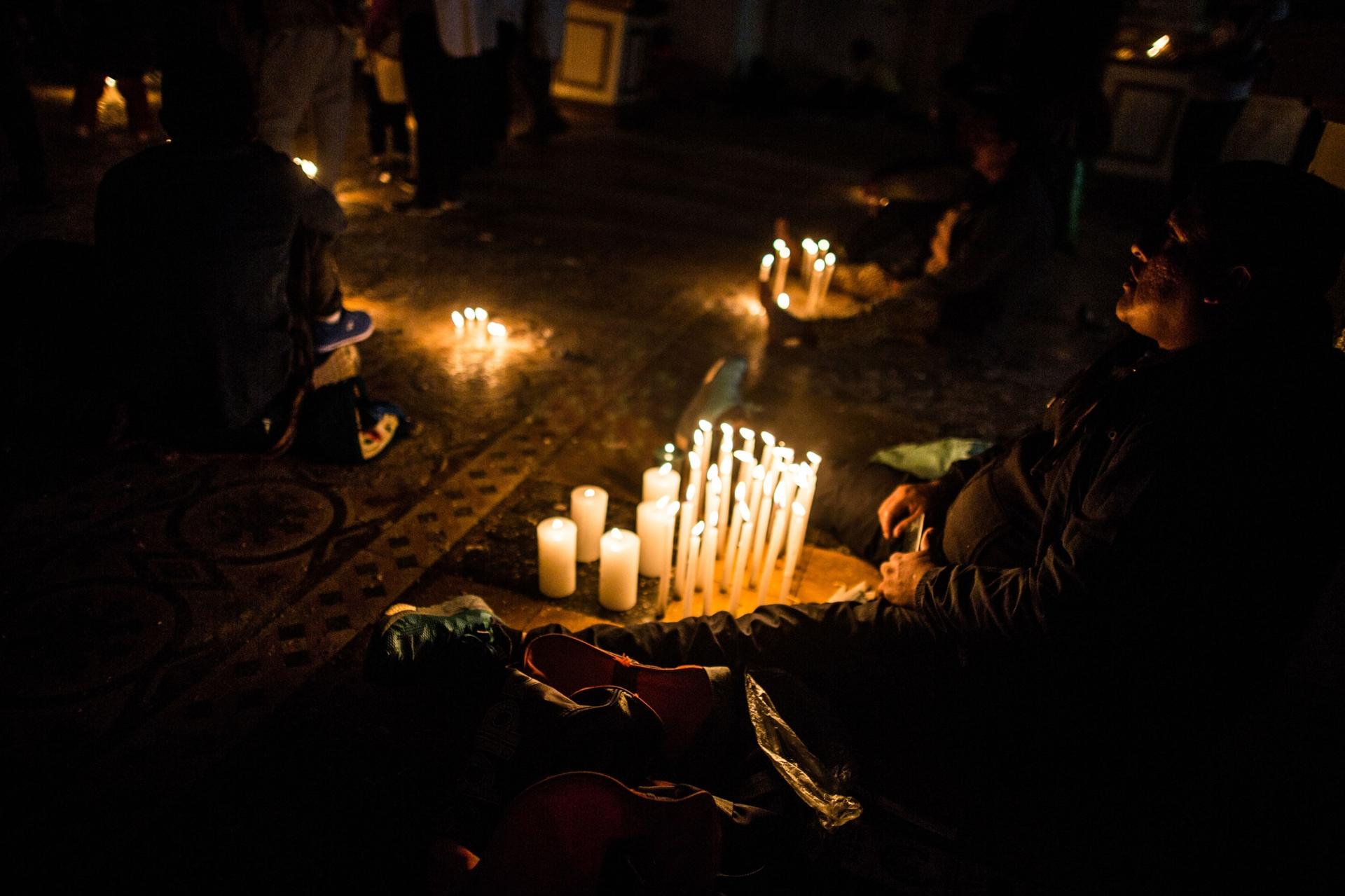 A person is shown sitting on the ground with their legs on either side of a display of several lit candles.