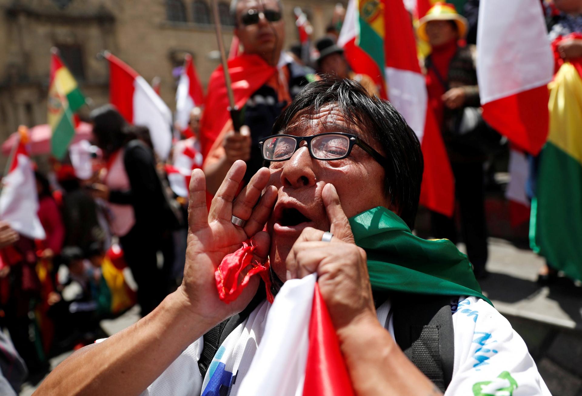 A demonstrator is shown with their hands around their mouth shouting and holding a Bolivian flag.