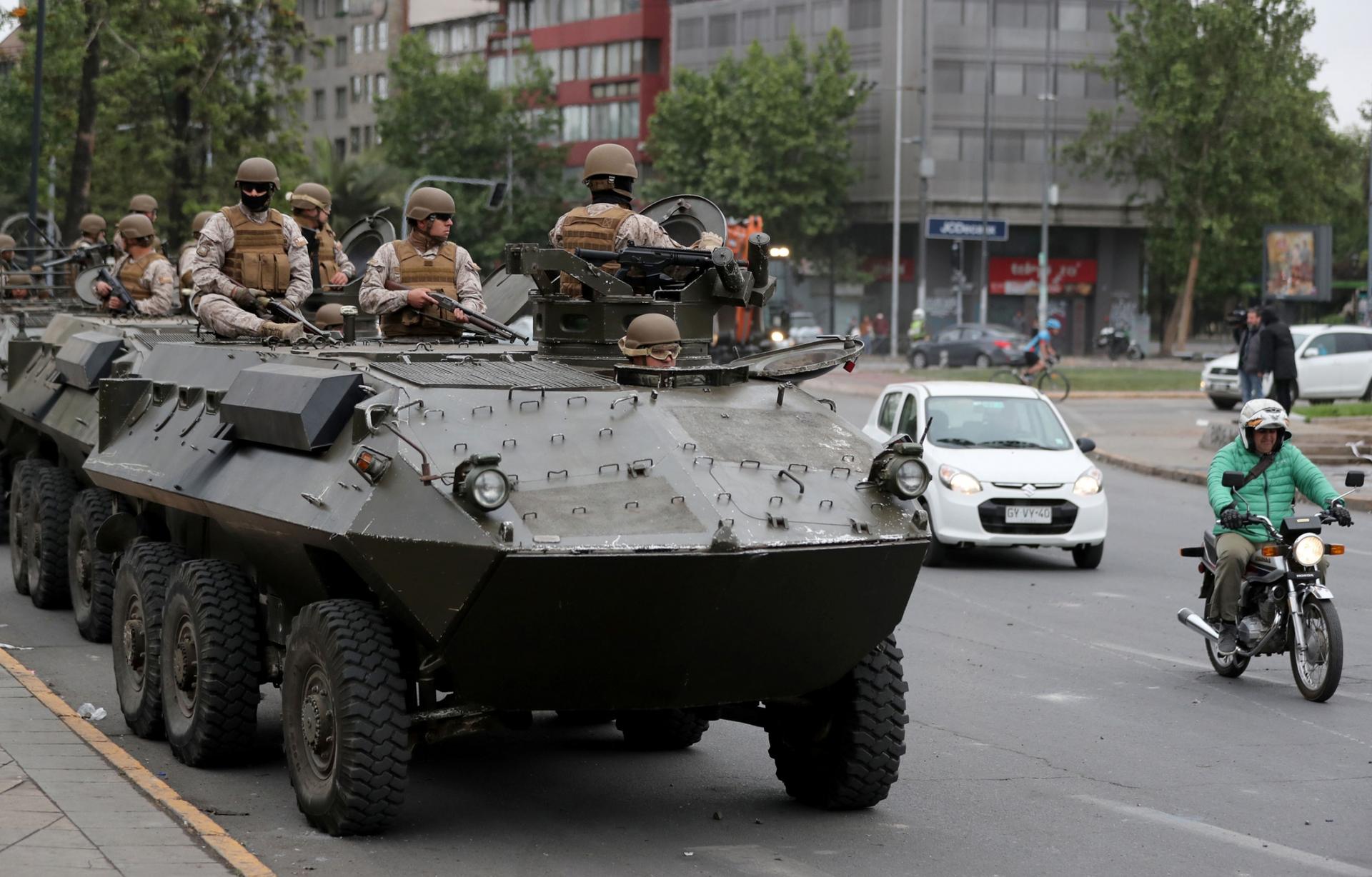 A line of armored transporter trucks are shown with armed soldiers riding on top and a man on a motorcycle passing by.