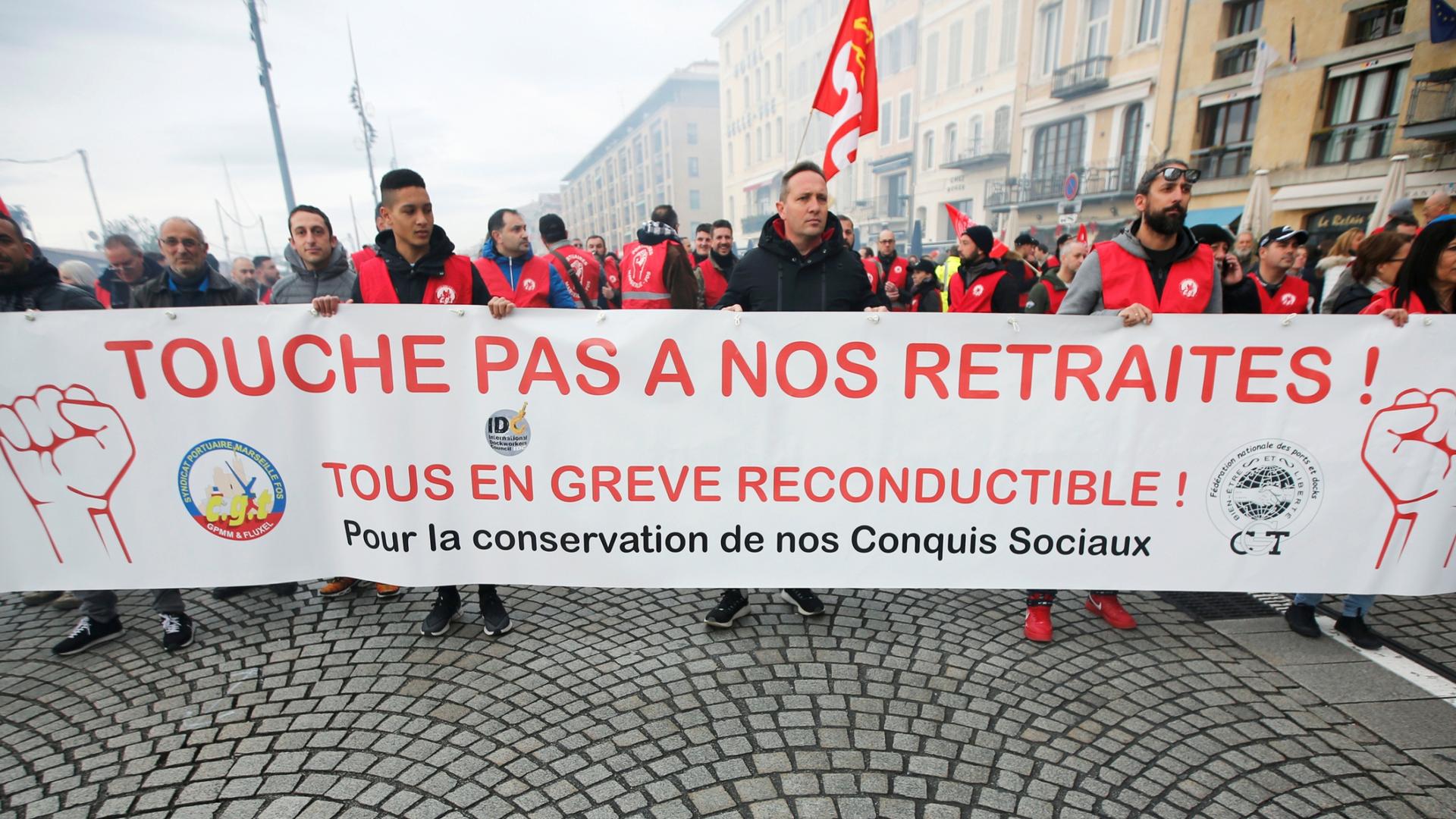 A large group of protesters are shown in the street holding a banner during a demonstration in Marseille. The banner reads, "Don't touch my pension."