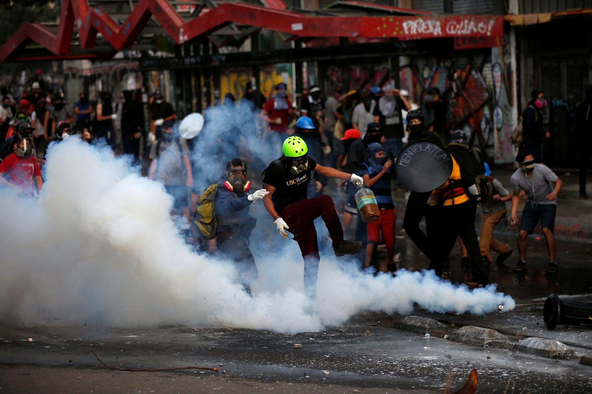 A goupr of demonstrators wearing gas masks are show surrounded by clouds of tear gas.