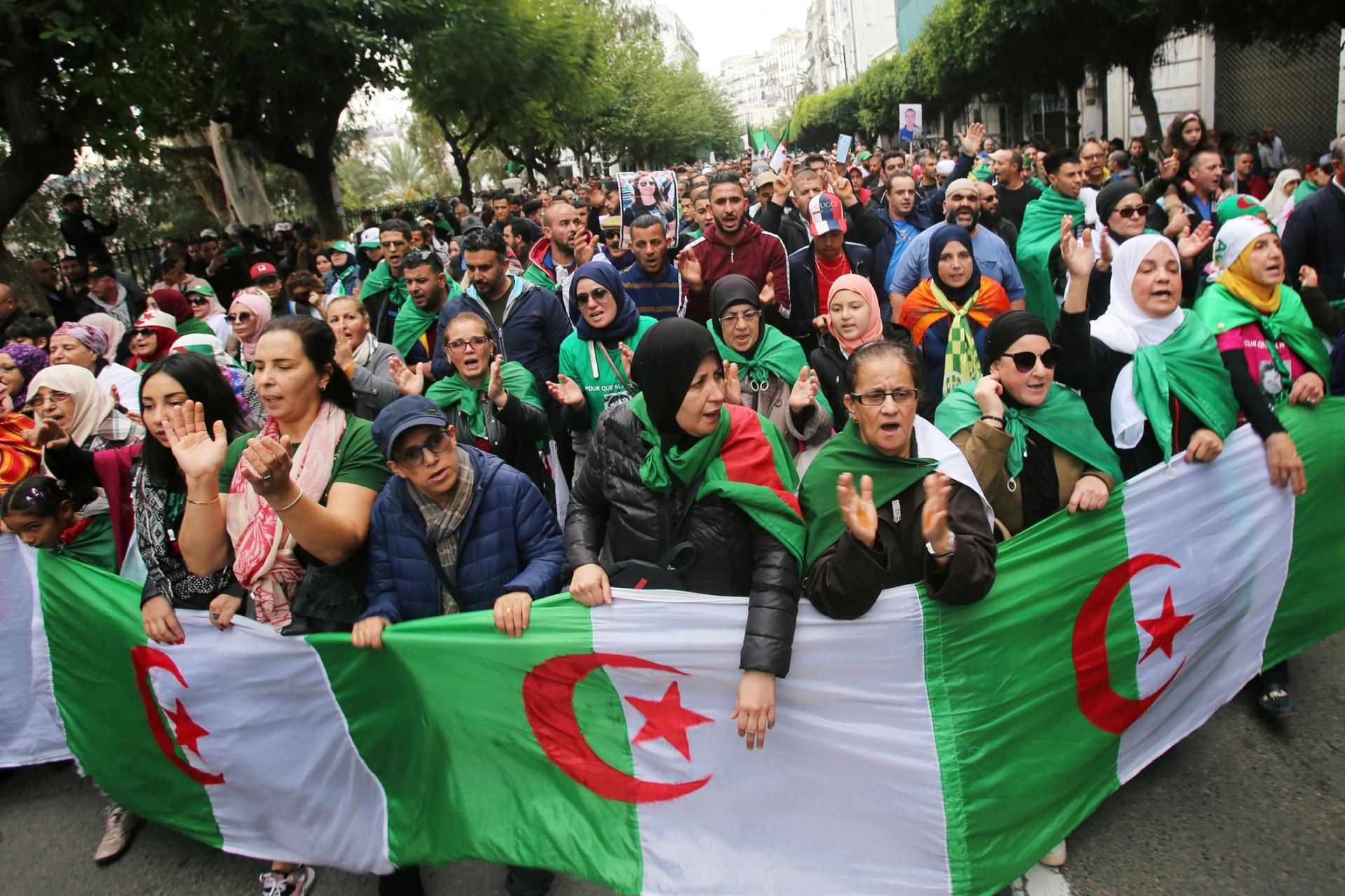 A large group of demonstrators are shown filling a street in Algiers with the Algerian flag in front of them.