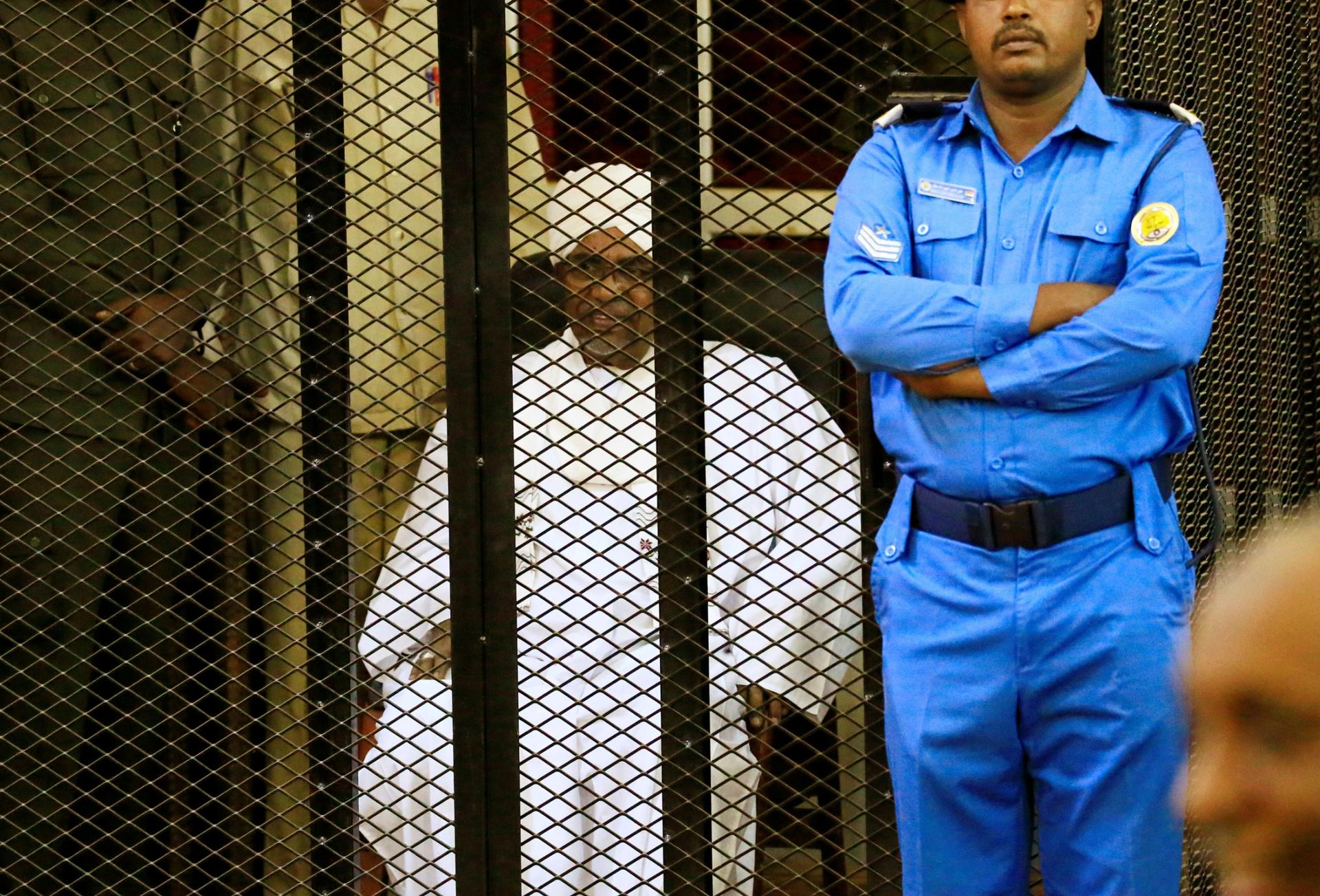 Sudanese former president Omar al-Bashir is shown sitting in all white and inside a metal cage with a guard standing beside it.