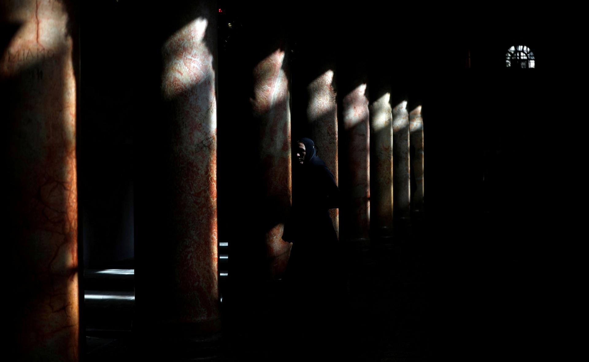 A worshipper is shown inside of the Church of the Nativity with very little light coming in and shinning on the tops of a row of marble columns.
