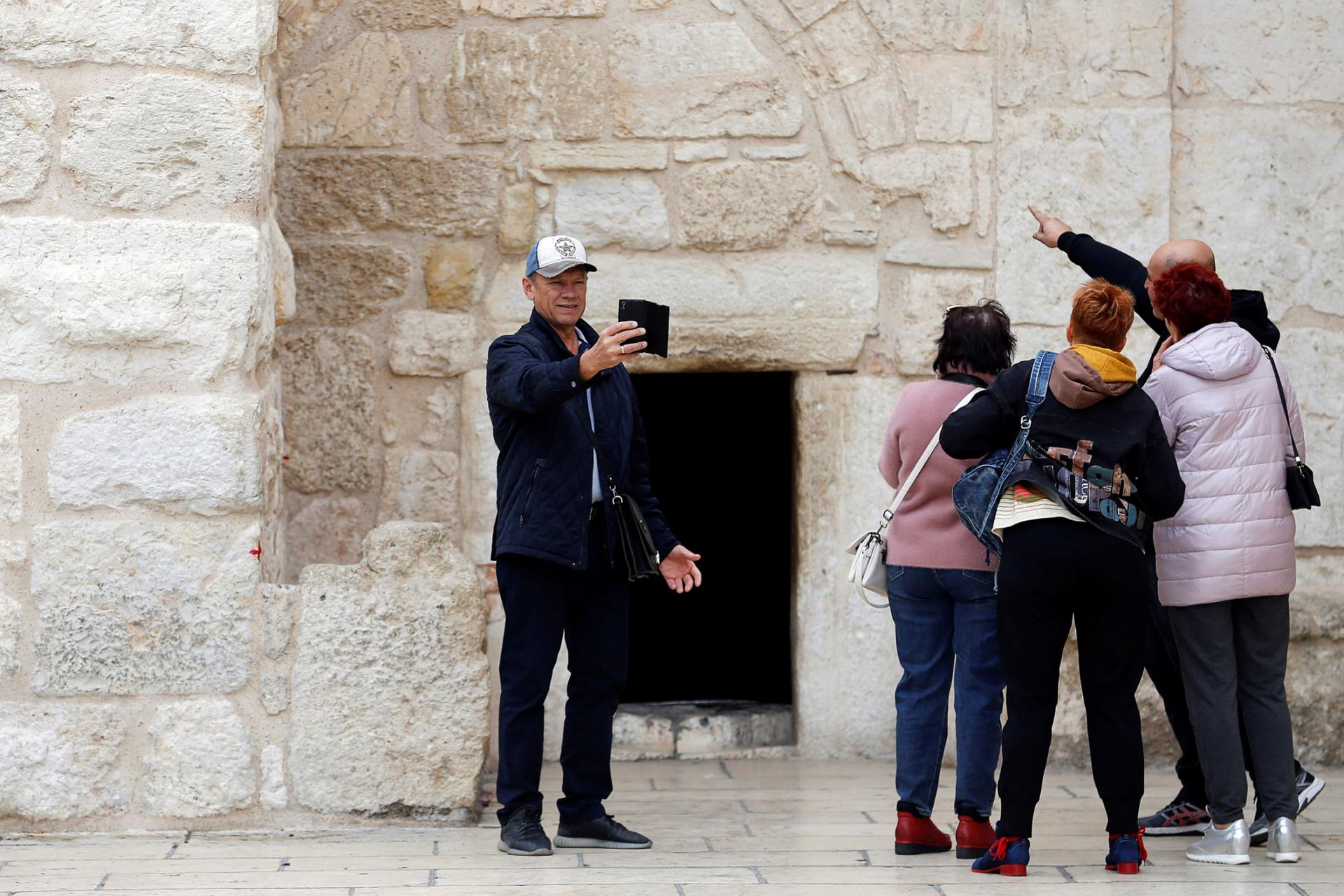 A tourist wearing a ball cap is shown with four other tourists taking a selfie outside the Church of the Nativity.