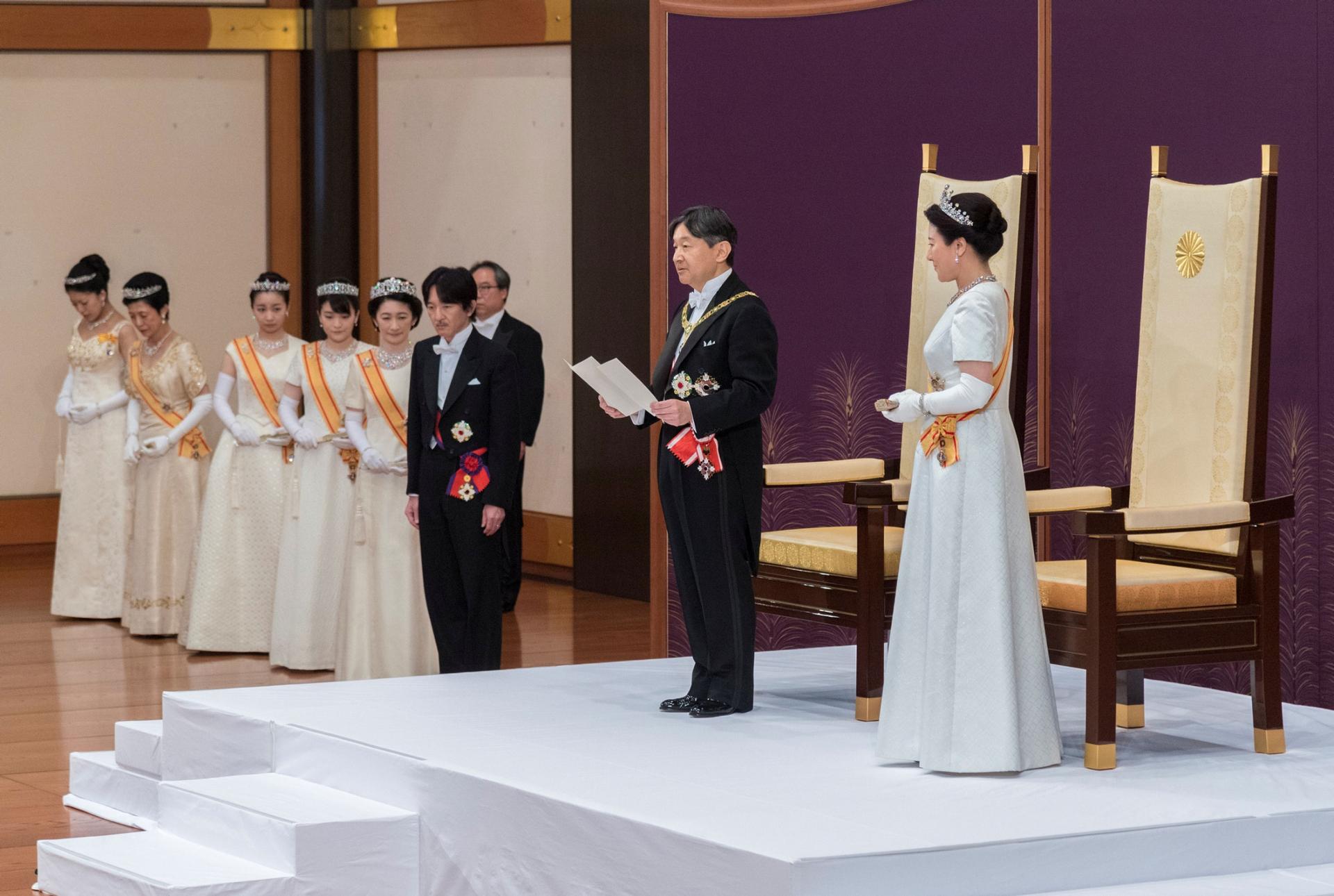 Japan's Emperor Naruhito, flanked by Empress Masako are shown standing on a white platform in front of tall-backed chairs.