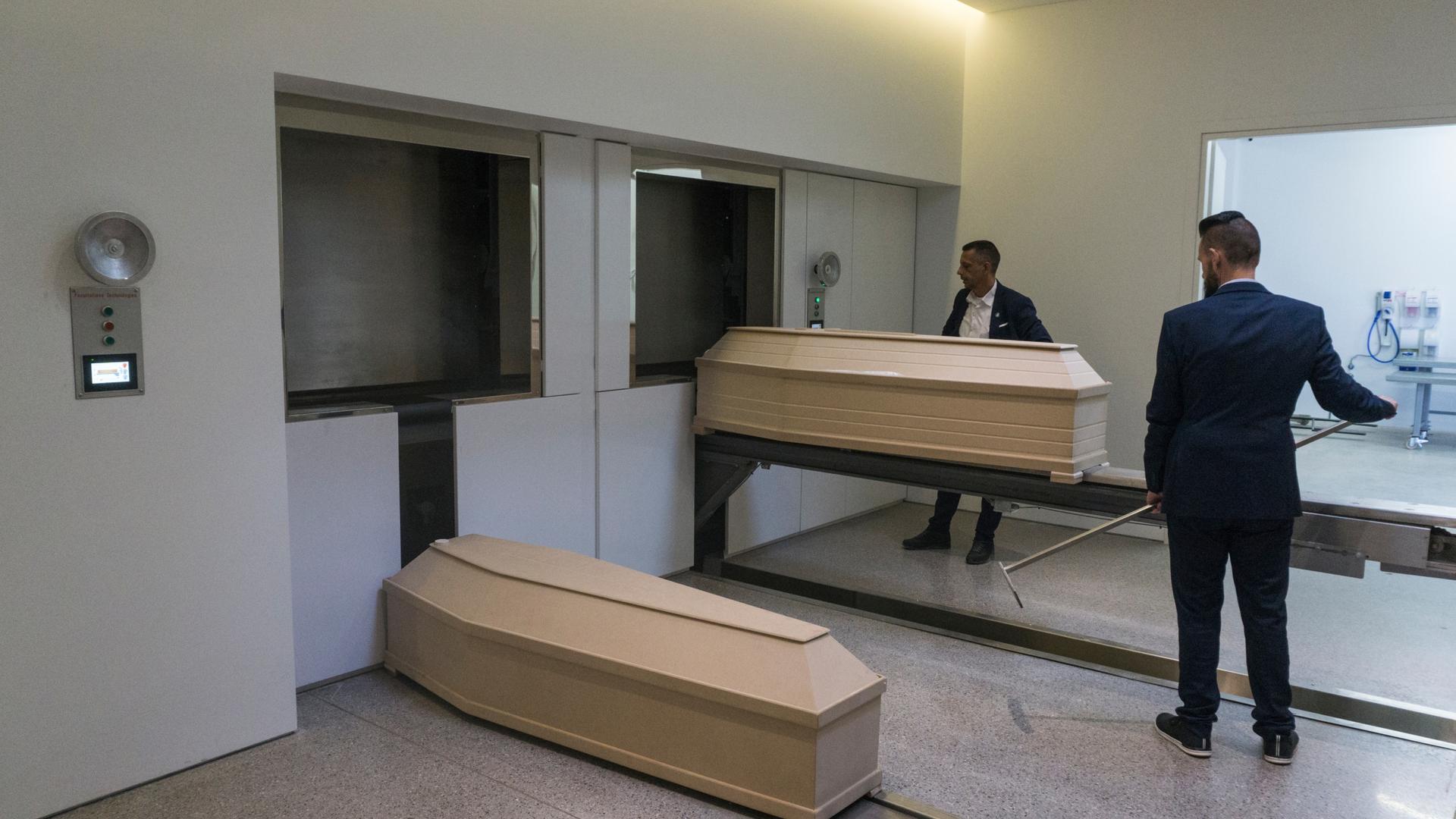 Two men in suits place beige-colored coffins in incinerators 