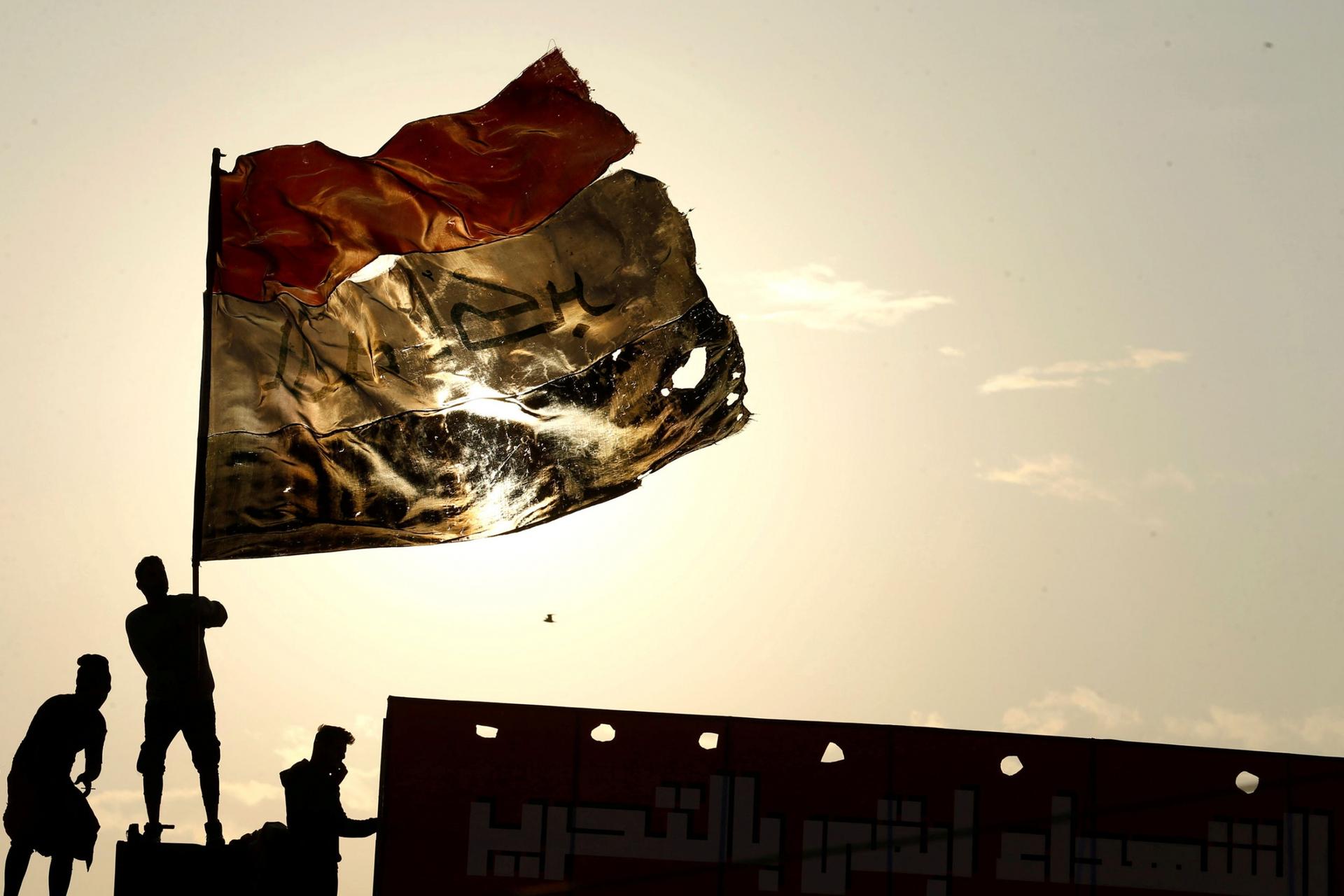 Three people are shown with one holding a large tattered Iraqi flag