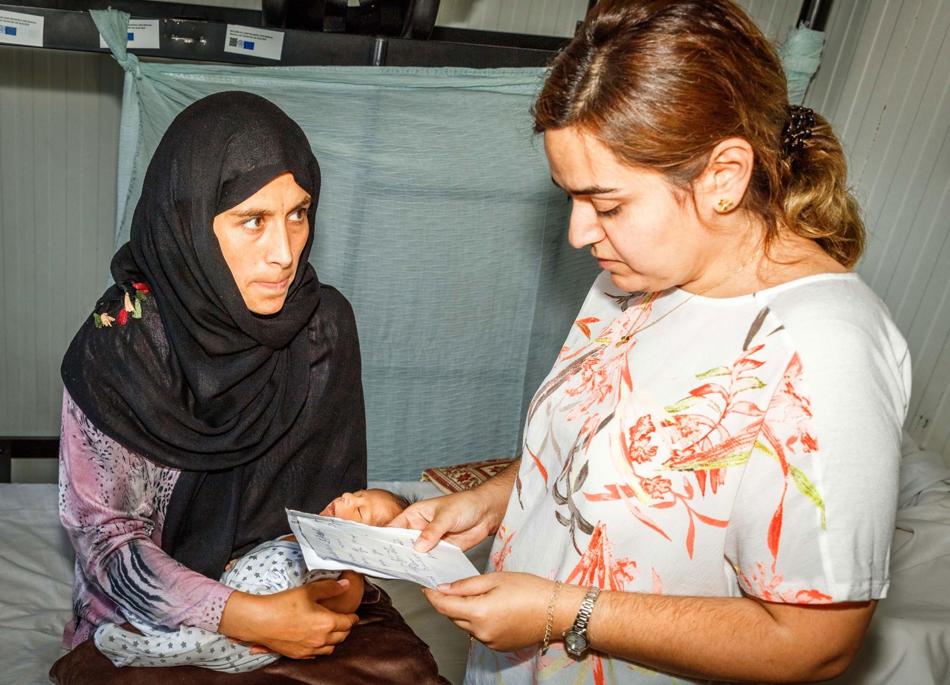 A woman is shown wearing a dark head scarf while holding an infant and looking at another woman holding papers.