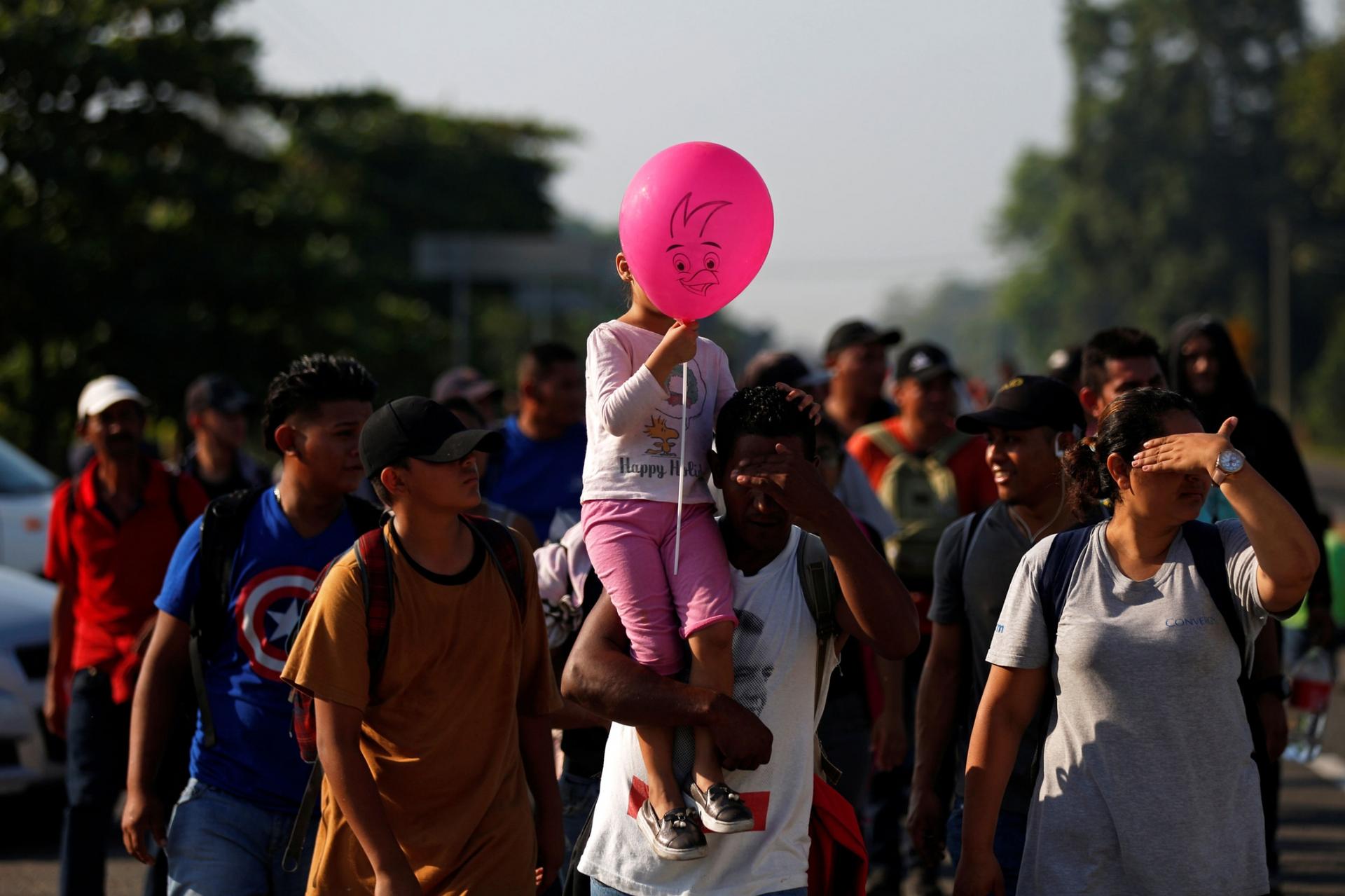 A large group of people are shown in the street walking and one person is carrying a young girl on their shoulder who has a pink balloon.