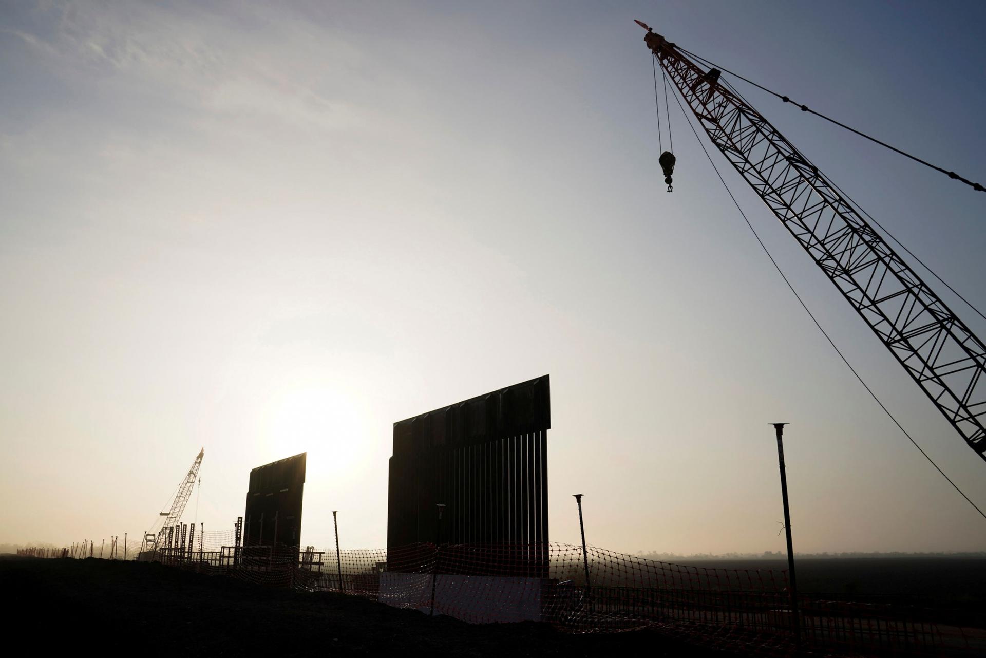 Two segments of the border wall are shown standing against a setting sun with a adjacent crane.