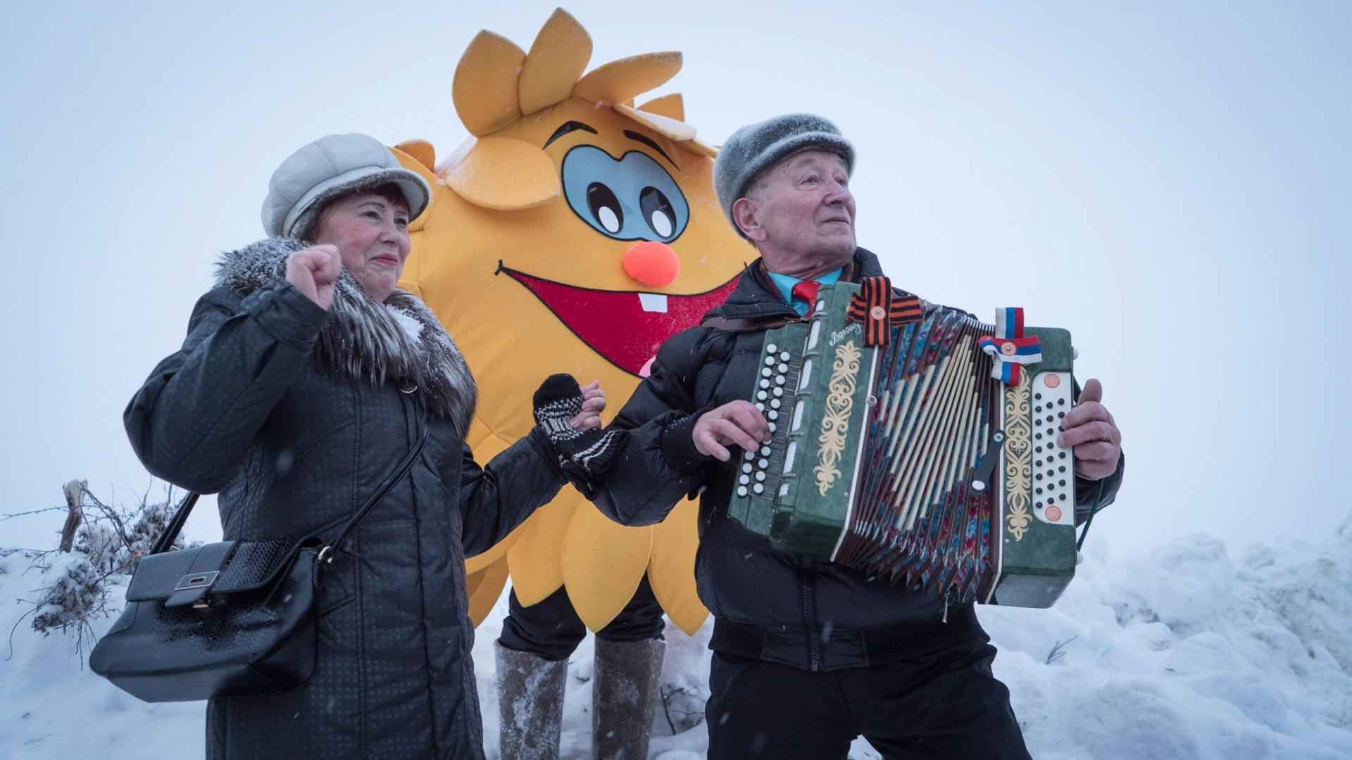 An older man is shown playing an accordion with a person dressed up in a sun costume stands behind him.