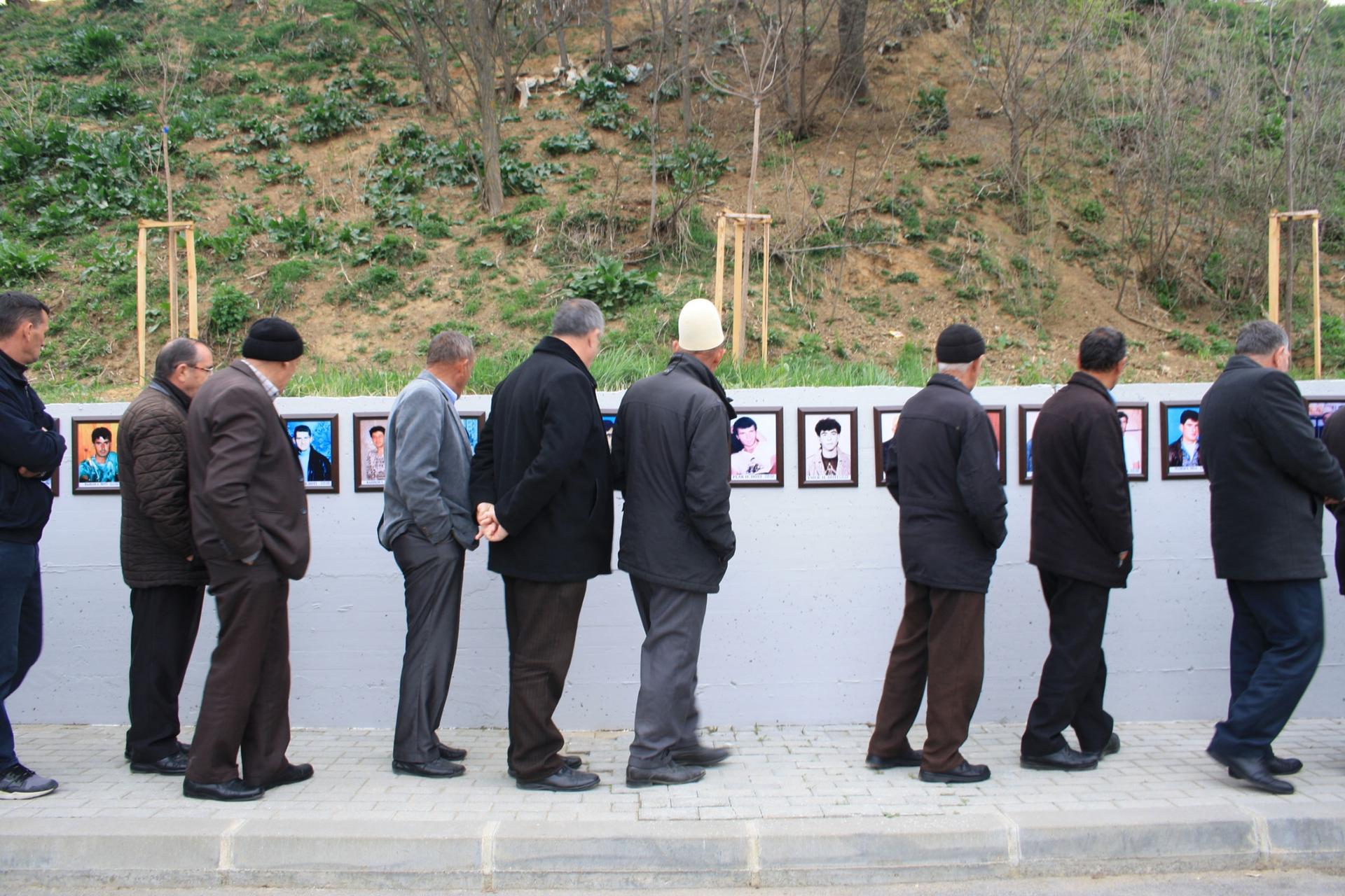 A line of men are shown standing next to a wall with photographs posted on it.