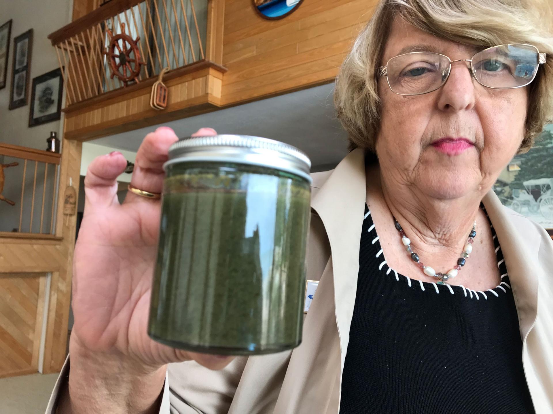 A woman is shown holding a glass jar and metal top filled with green algae water.