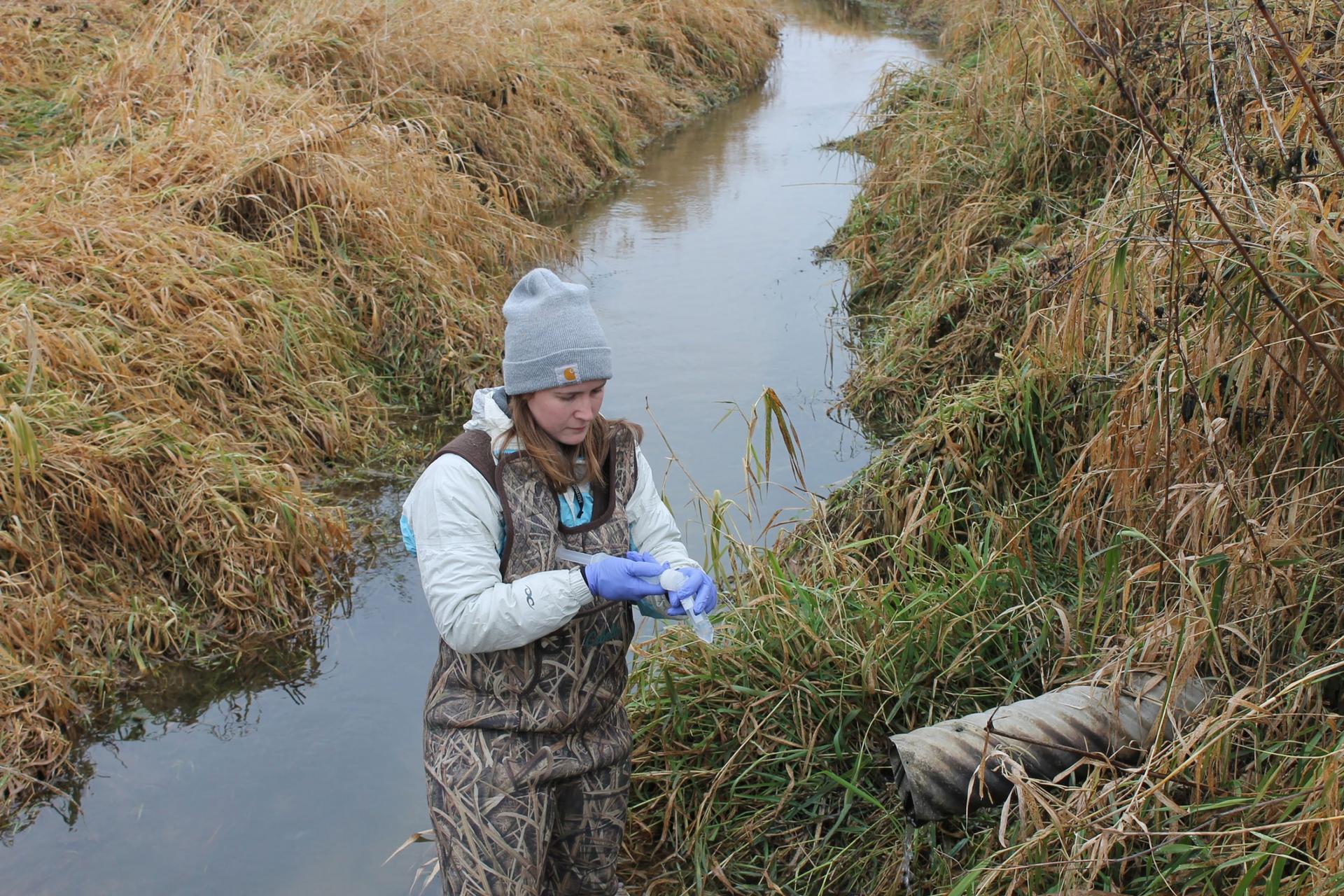 A woman is shown standing in a watery ditch and wearing camoflauge overalls.